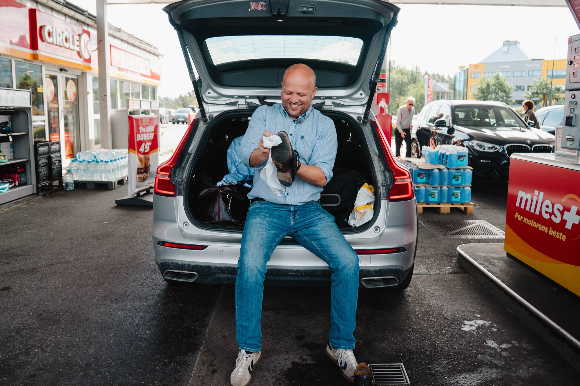 Mann sitter i bagasjerommet på en bil på en bensinstasjon og pusser en sko. (Bildebeskrivelsen er laget av en KI-tjeneste)