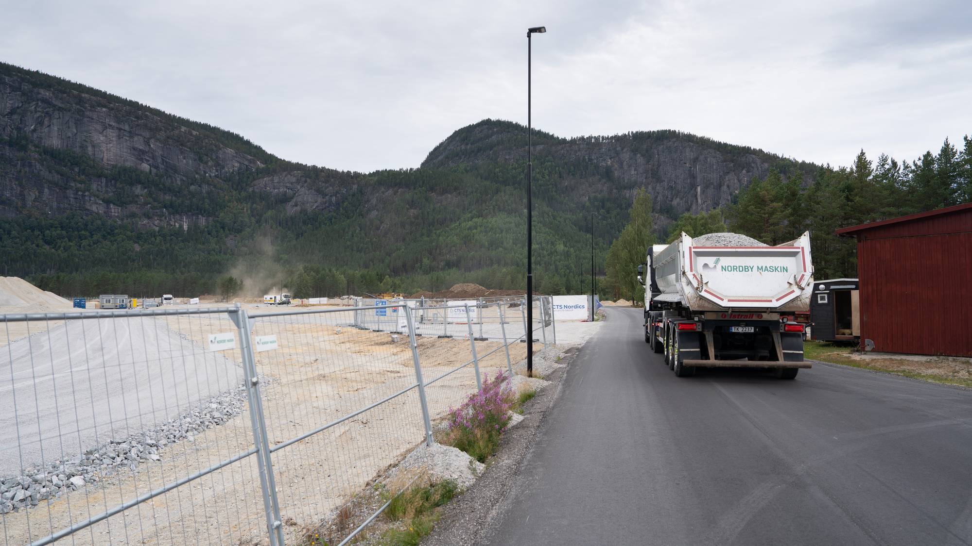 Lastebil kjører på vei ved anleggsområde med fjell i bakgrunnen. (Bildebeskrivelsen er laget av en KI-tjeneste)