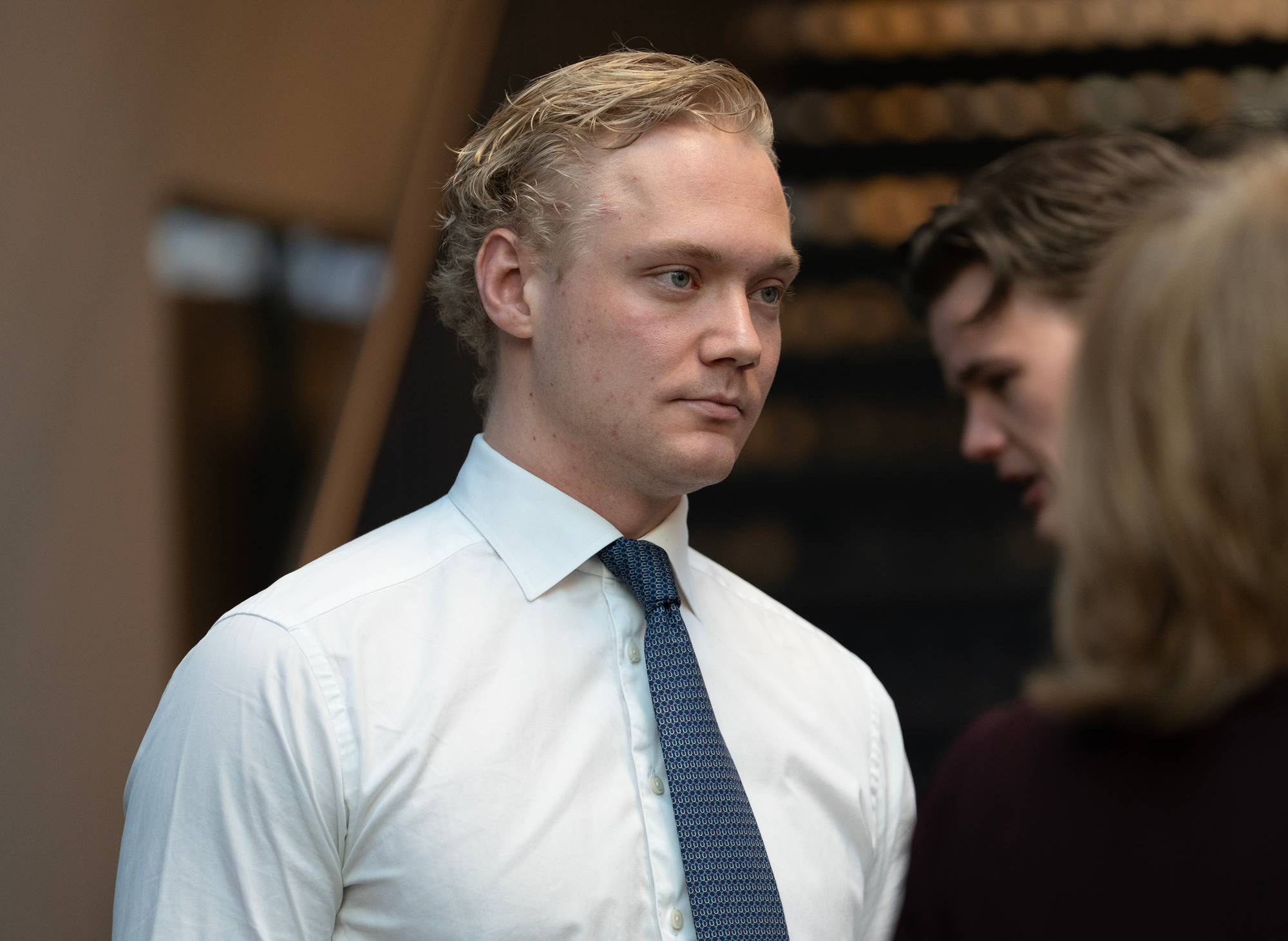 En ung mann i hvit skjorte og slips ser mot siden. Bak ham står to personer. Mannen har blondt hår. (Bildebeskrivelsen er laget av en KI-tjeneste)