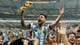 AP En fotballspiller i Argentinas drakt hever FIFA verdensmesterskapstroféet. Han er omringet av feirende supportere. Arenaen har en stor kapasitet, og mange tilskuere er til stede. Stemningen er festlig, med folk som tar bilder og jubler.