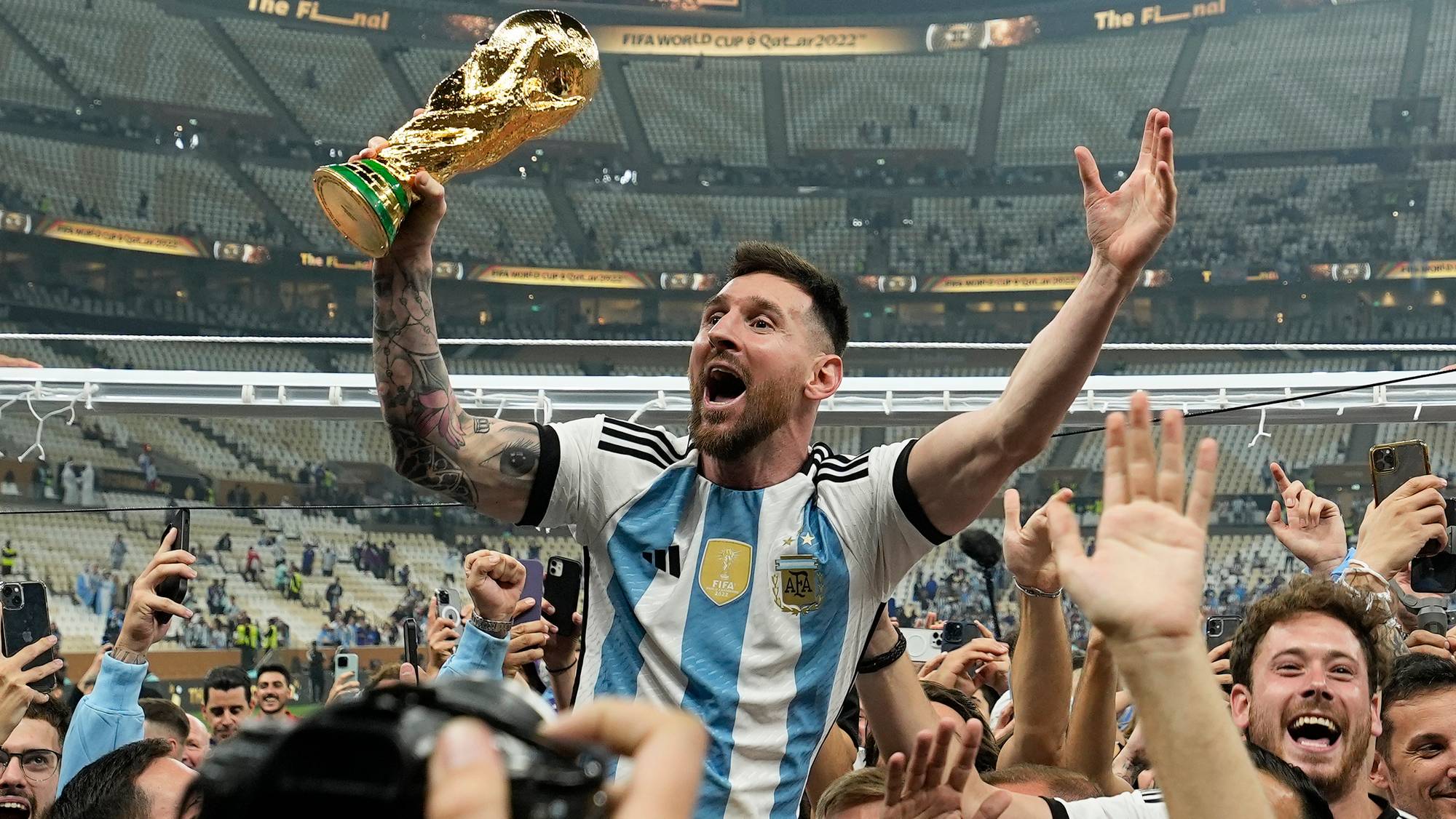 En fotballspiller i Argentinas drakt hever FIFA verdensmesterskapstroféet. Han er omringet av feirende supportere. Arenaen har en stor kapasitet, og mange tilskuere er til stede. Stemningen er festlig, med folk som tar bilder og jubler.