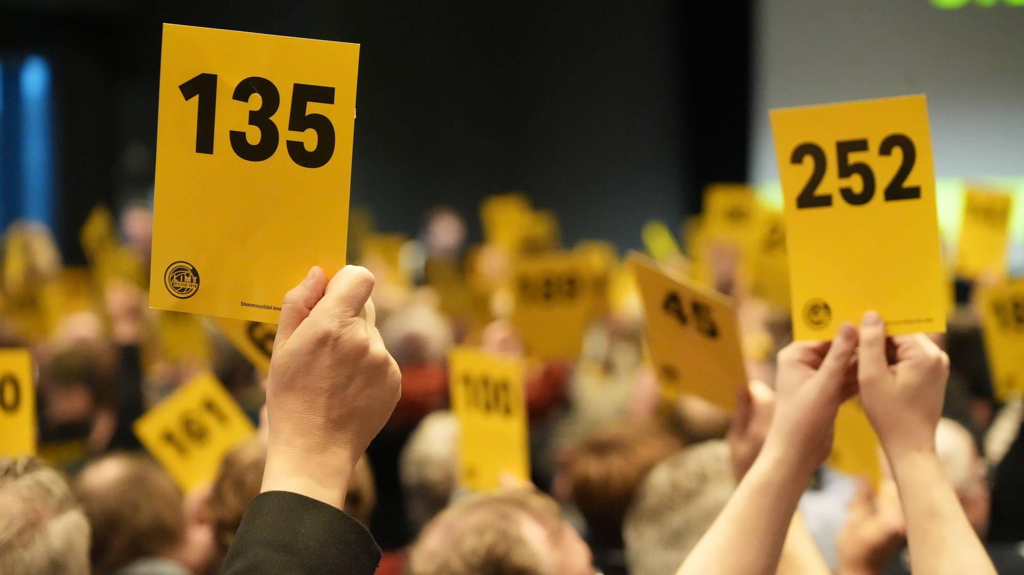 Folk holder opp gule stemmesedler med nummer i en forsamling. (Bildebeskrivelsen er laget av en KI-tjeneste)
