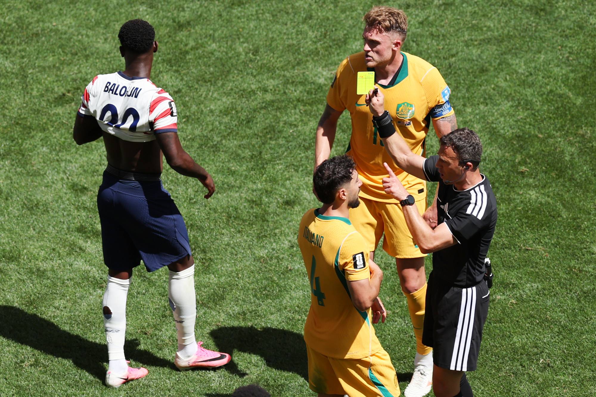Dommer gir gult kort til spiller. Dommeren holder opp et gult kort foran en spiller i gult. En annen spiller i gult står ved siden av. En spiller med nummer 20 på ryggen, med navnet BALOJUIN, står vendt bort fra kameraet. Alle befinner seg på en grønn gressmatte. (Bildebeskrivelsen er laget av en KI-tjeneste)