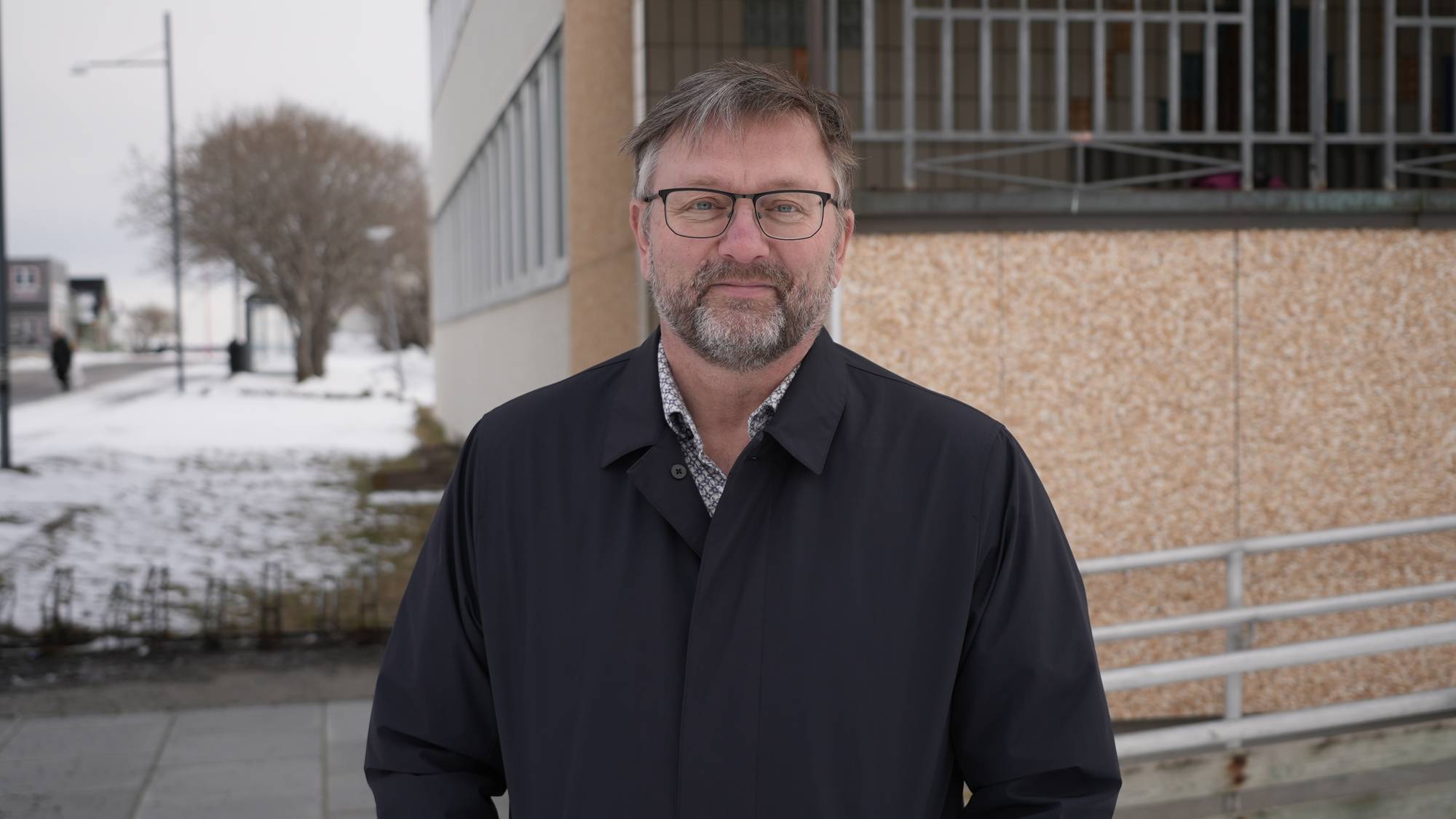 Middelaldrende mann med briller og mørk jakke står utendørs ved en bygning. (Bildebeskrivelsen er laget av en KI-tjeneste)