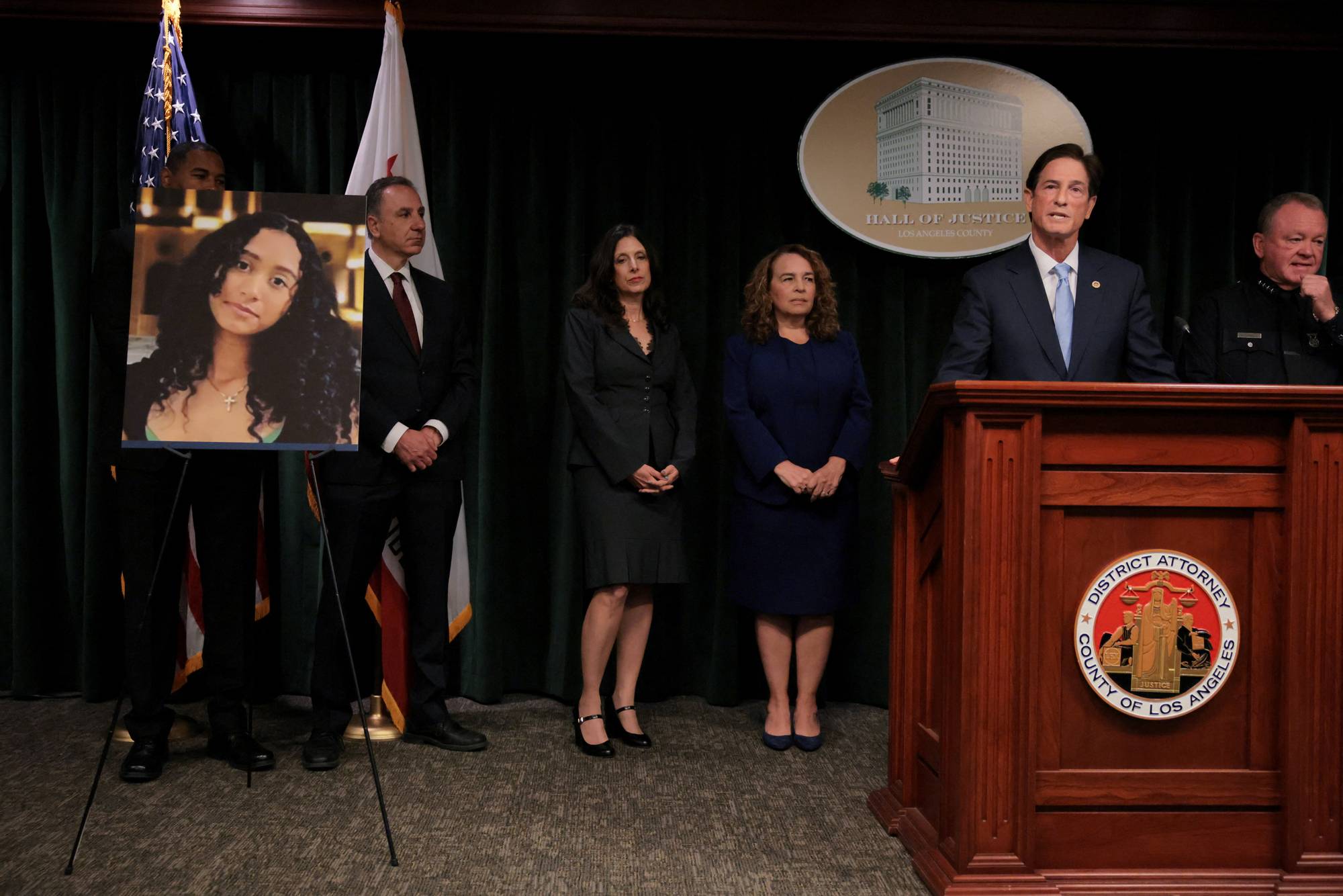 En mann i dress taler bak et podium med logo for Los Angeles County District Attorney. Bak ham står fire personer og et bilde av en ung kvinne på et staffeli. (Bildebeskrivelsen er laget av en KI-tjeneste)
