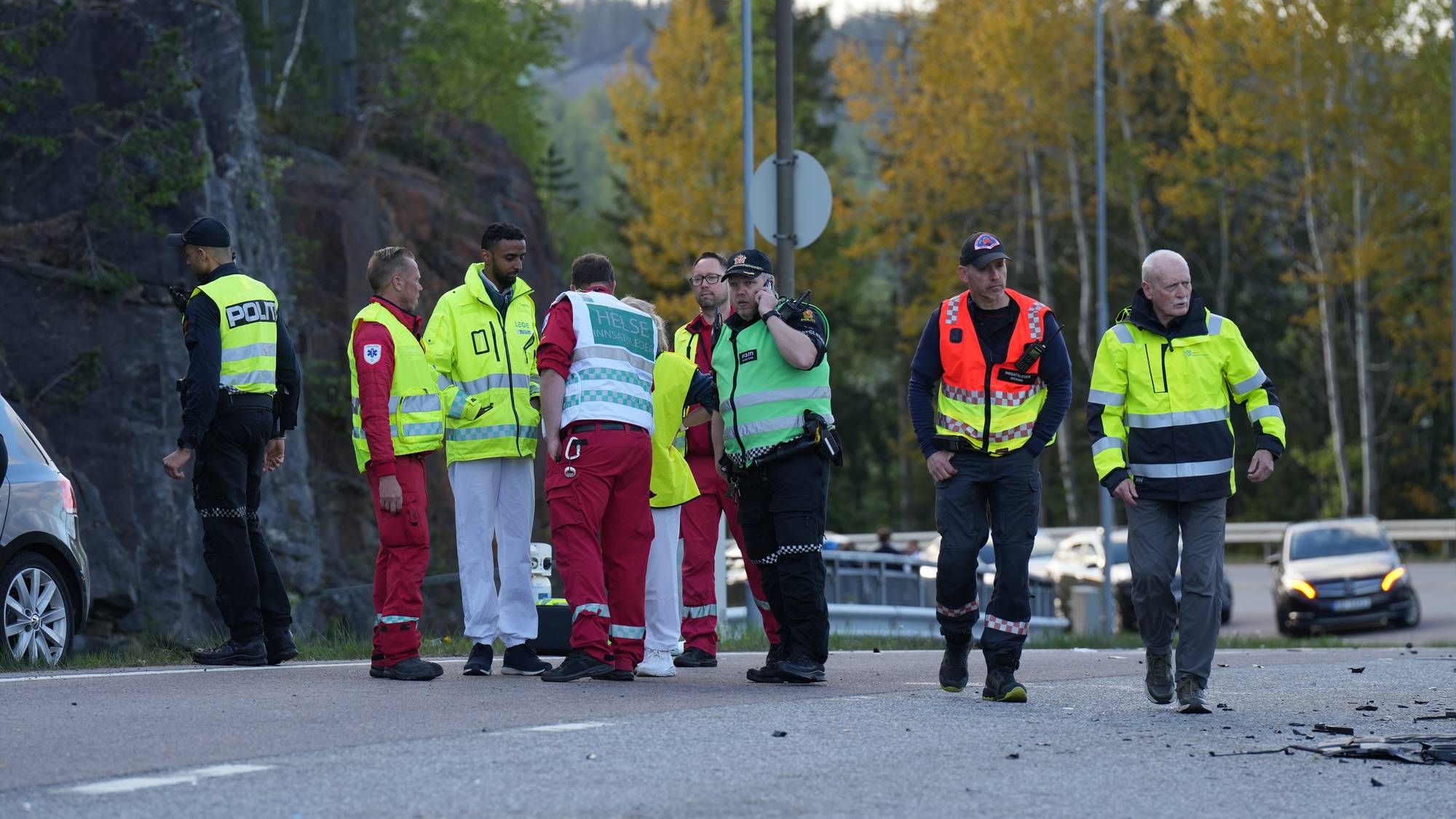 Personell fra politi og helsevesen er samlet på en vei ved et skogholt. De står i en gruppe og ser ut til å være i samtale. En mann snakker i en mobiltelefon, mens en annen snakker med noen i gruppen. Flere personer er iført gule og oransje refleksvester. Bakgrunnen viser trær og en fjellside. (Bildebeskrivelsen er laget av en KI-tjeneste)