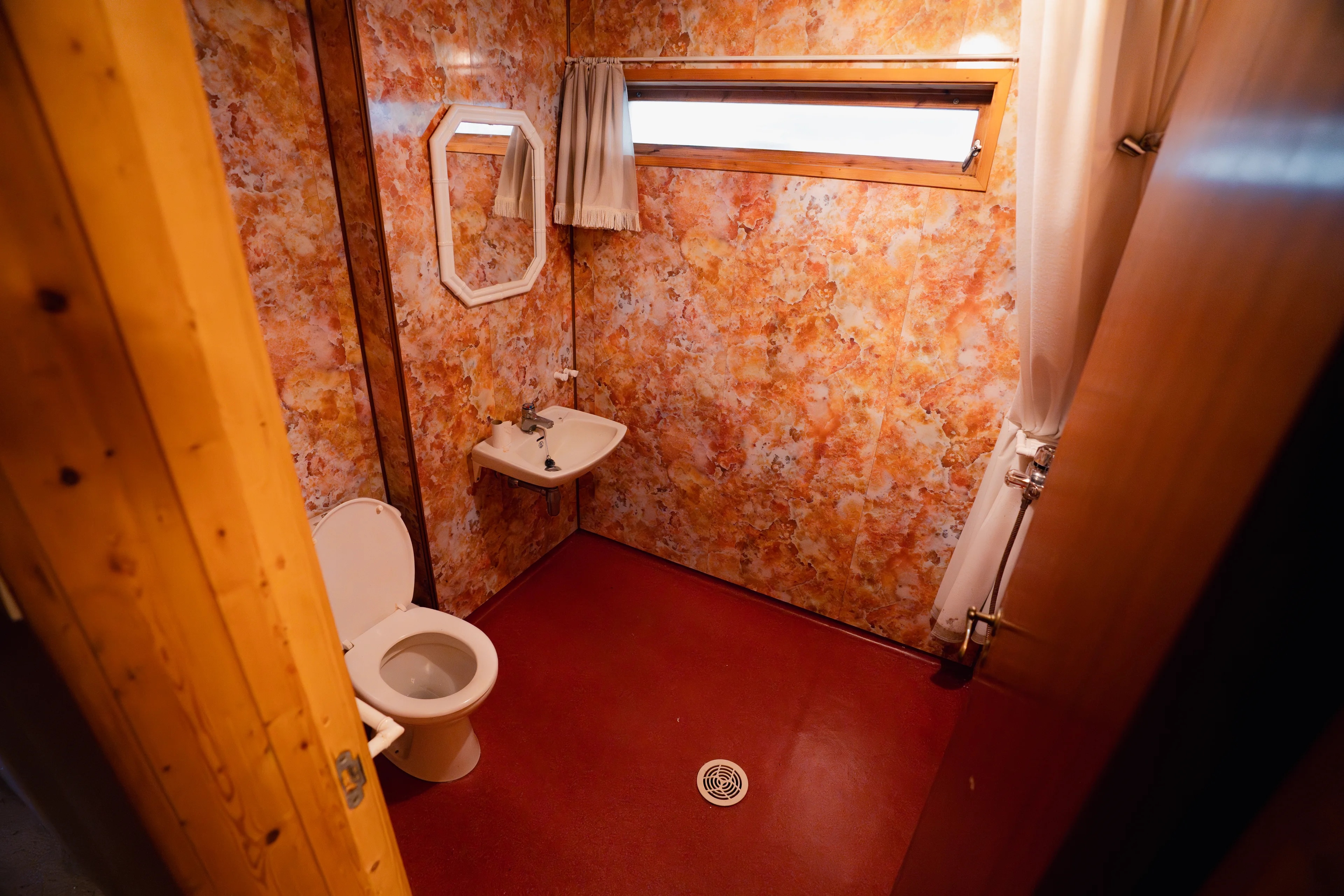 Bad med oransje marmorerte vegger, rødt gulv, toalett, servant, speil og et avlangt vindu. (Bildebeskrivelsen er laget av en KI-tjeneste)