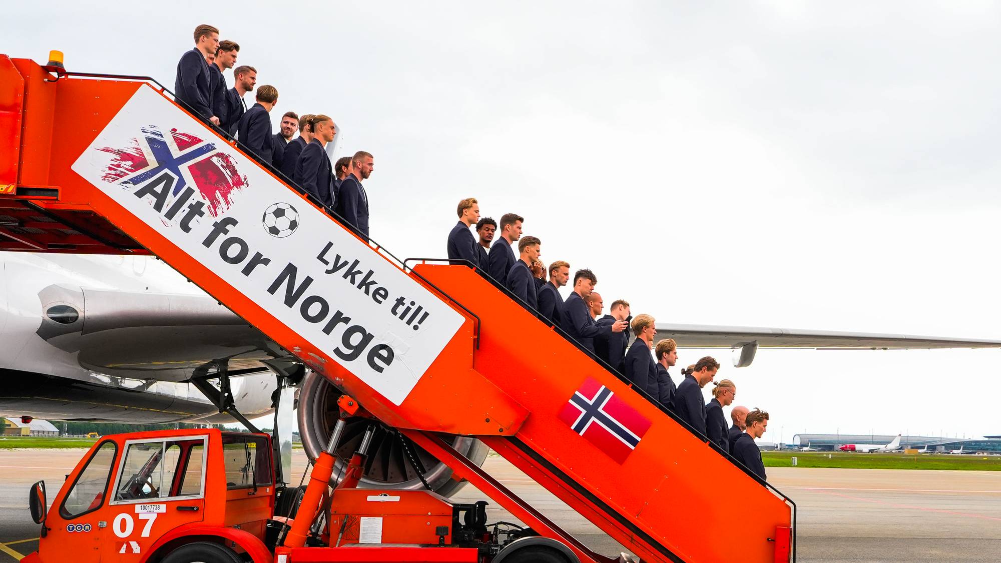 En gruppe menn i mørke dresser står på en oransje flytrapp. Teksten "Alt for Norge Lykke til!" og et norsk flagg er synlig på trappen. Mennene er på vei mot et fly, og en bil står parkert under trappen. (Bildebeskrivelsen er laget av en KI-tjeneste)