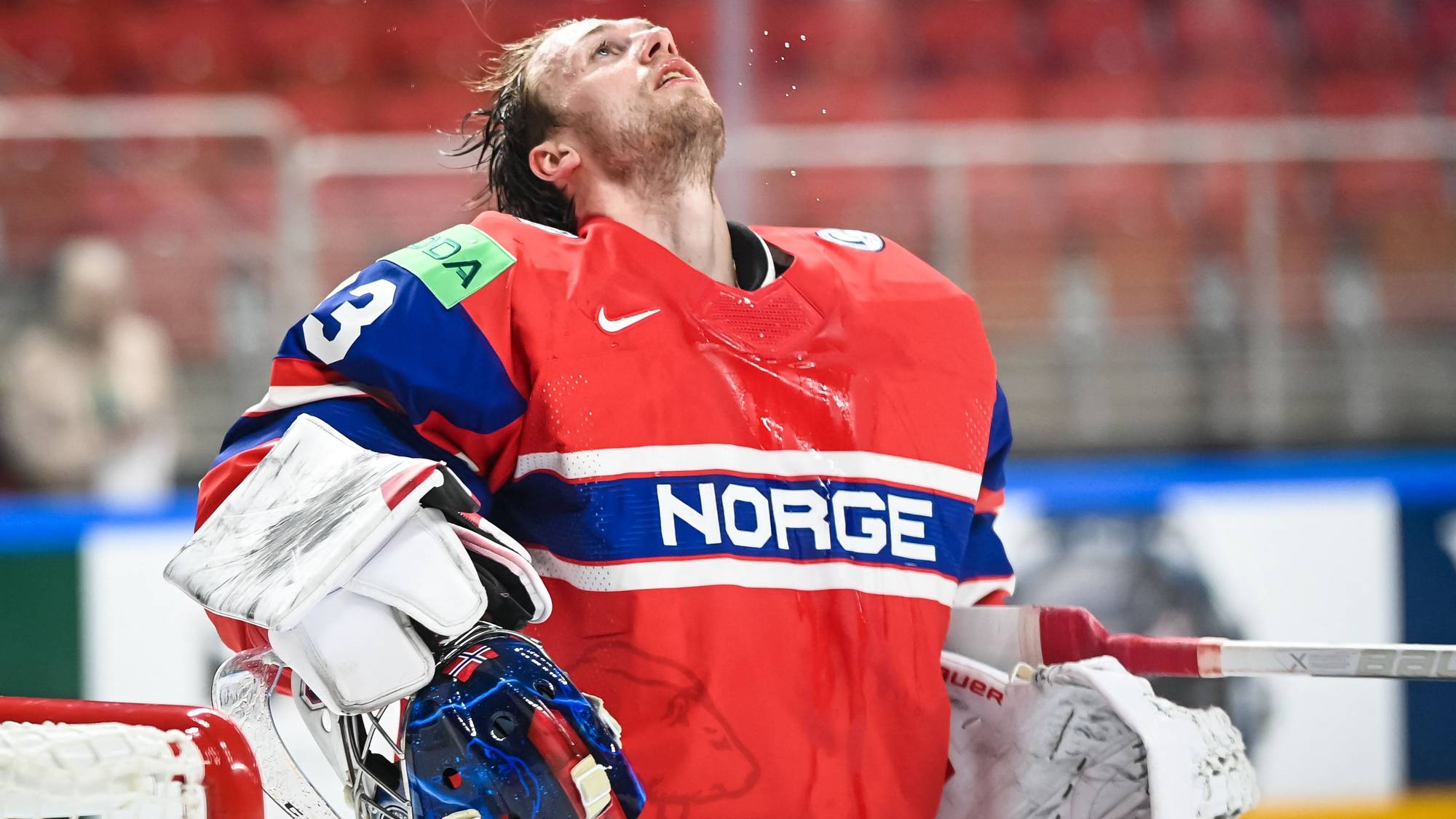 En ishockeymålvakt i Norges drakt med nummer 3 på ryggen ser oppover. Målvakten har en hjelm med blått og svart mønster og store hansker. I bakgrunnen er det røde tribuner. (Bildebeskrivelsen er laget av en KI-tjeneste)