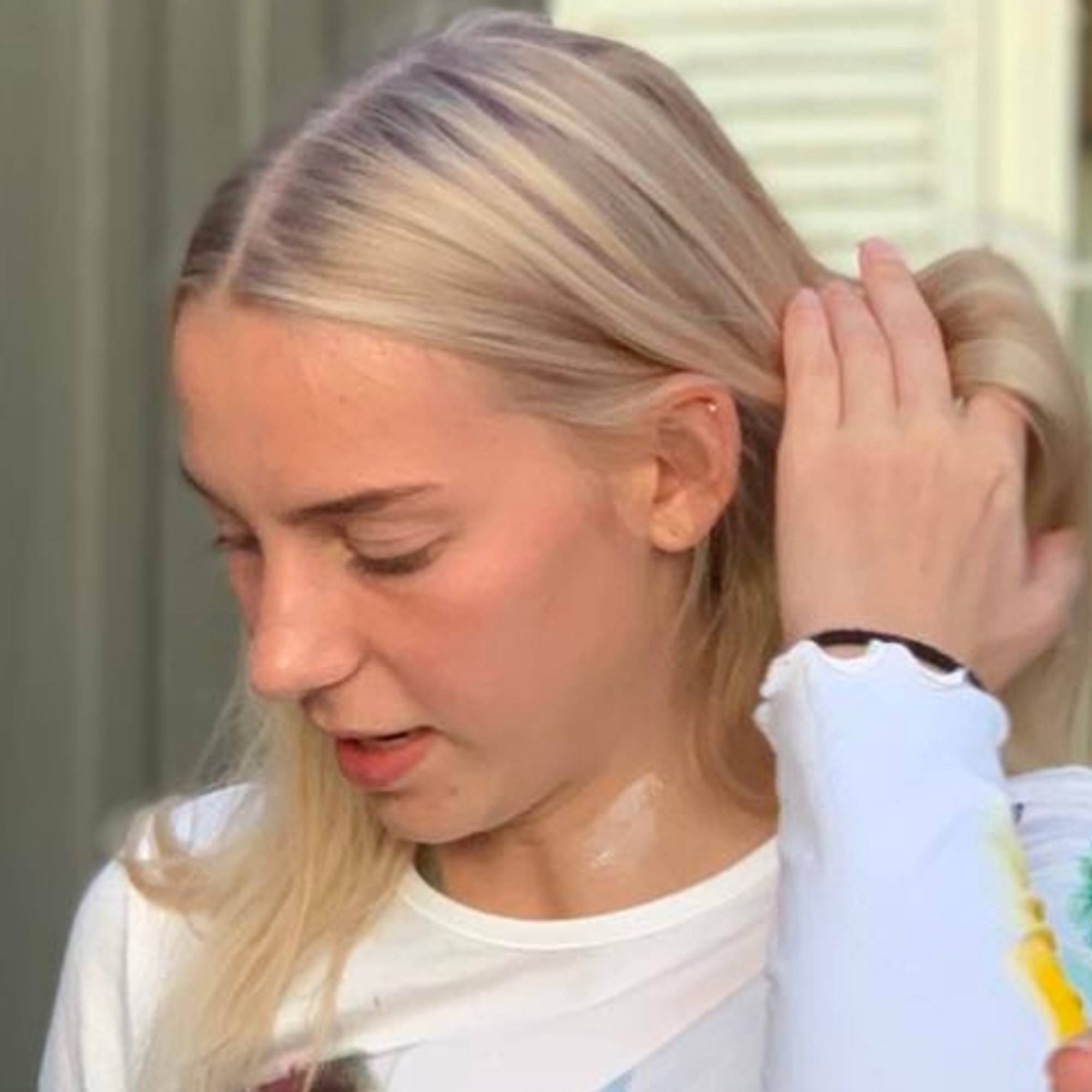Cathrine Michelsen med fjær og farger på kinnet. Ser ned og tar det blonde håret bakover. 