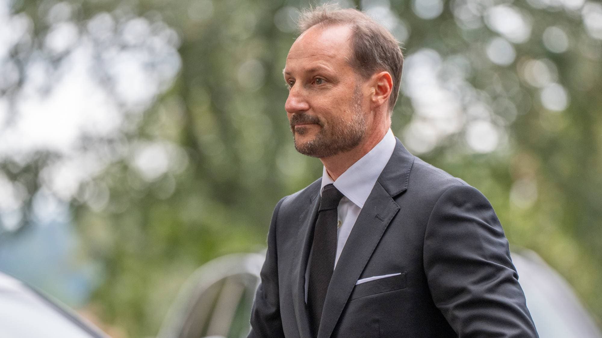 Kronprins Haakon i dress og slips, fotografert utendørs med trær i bakgrunnen. (Bildebeskrivelsen er laget av en KI-tjeneste)