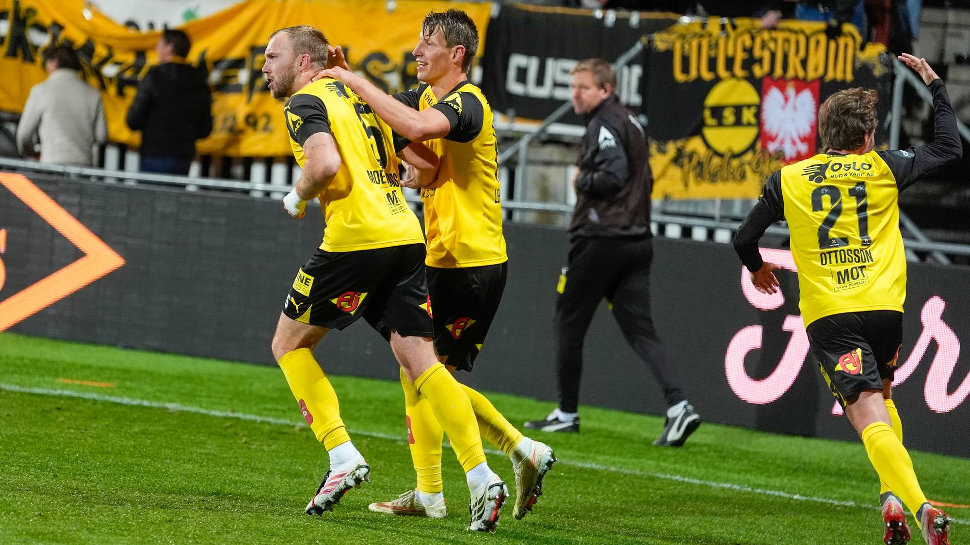 Fotballspillere fra Lillestrøm feirer mål i gule drakter på en fotballbane. (Bildebeskrivelsen er laget av en KI-tjeneste)