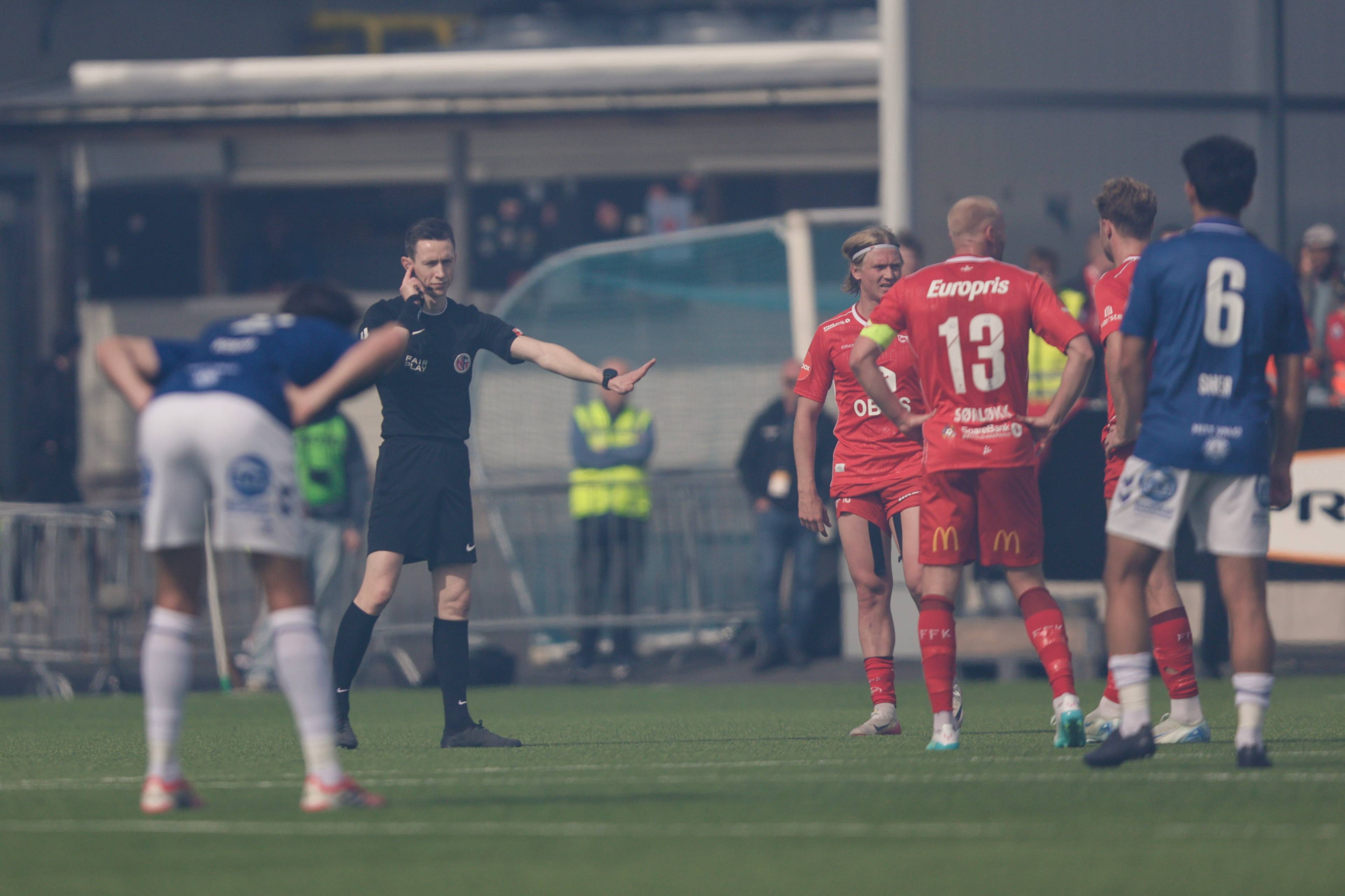 En dommer i svart uniform vinker spillere til side under en fotballkamp. Flere spillere i røde og blå drakter står på gressmatten. I bakgrunnen skimtes tribuneanlegg og andre personer. (Bildebeskrivelsen er laget av en KI-tjeneste)