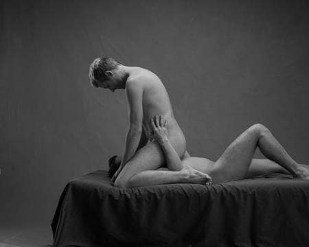 En blond mann kneler over en mann med mørkt hår som ligger på ryggen. Han sitter over ansiktet hans med beina spredt. De simulerer oralsex. Begge er nakne, men kjønnsorganene er ikke synlige.