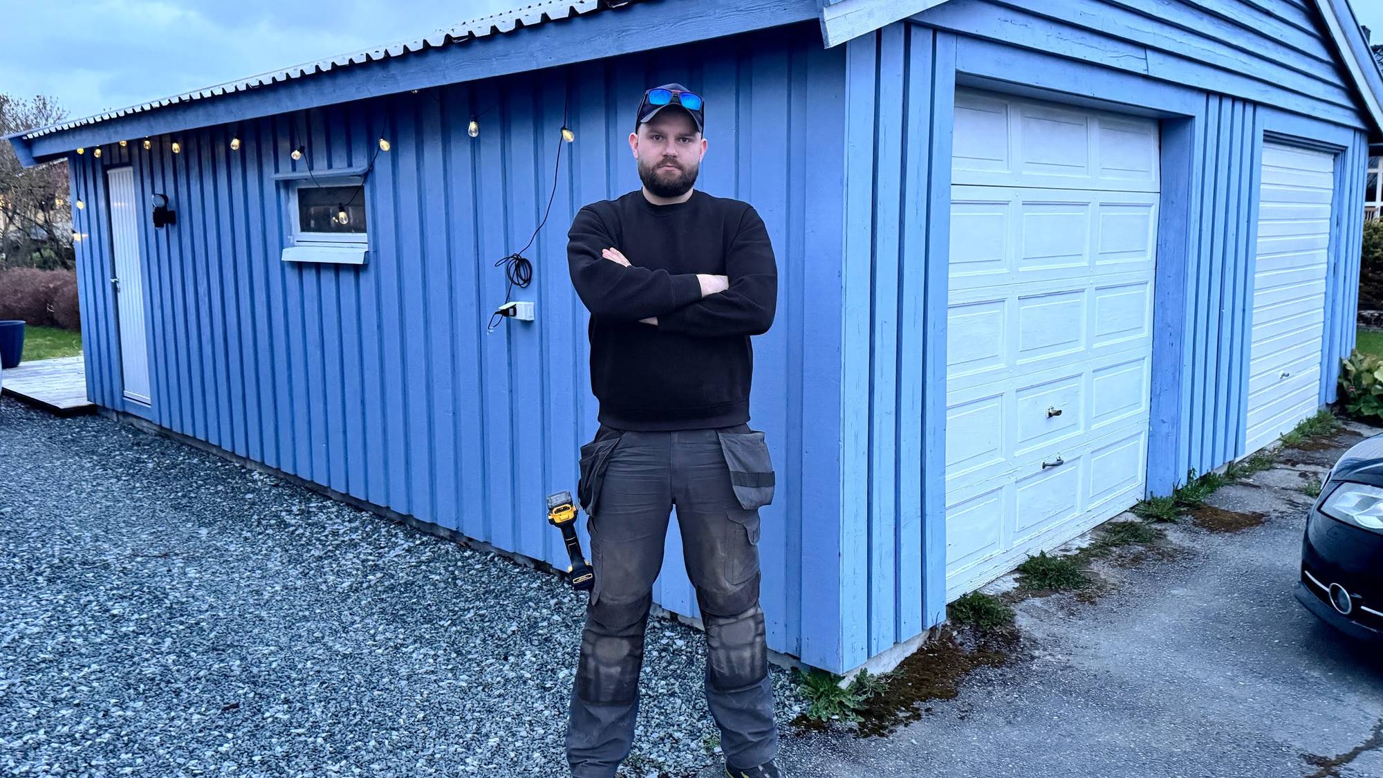 Mann med caps, svart genser foran blå dobbel garasje.