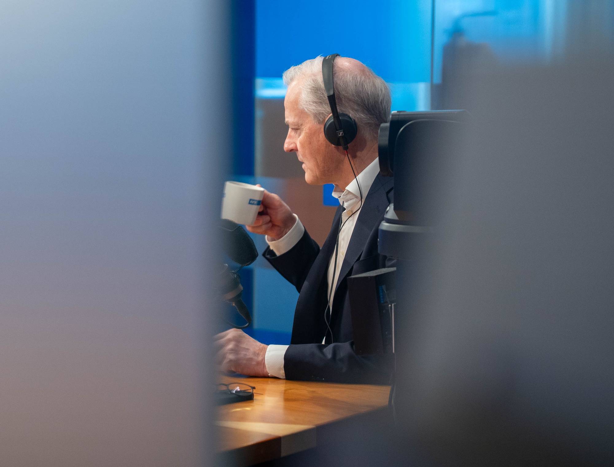 En eldre mann med hodetelefoner sitter ved et bord og holder en kopp. Han har på seg mørk dress og hvit skjorte. En mikrofon står på bordet foran ham. Bakgrunnen er blå. (Bildebeskrivelsen er laget av en KI-tjeneste)