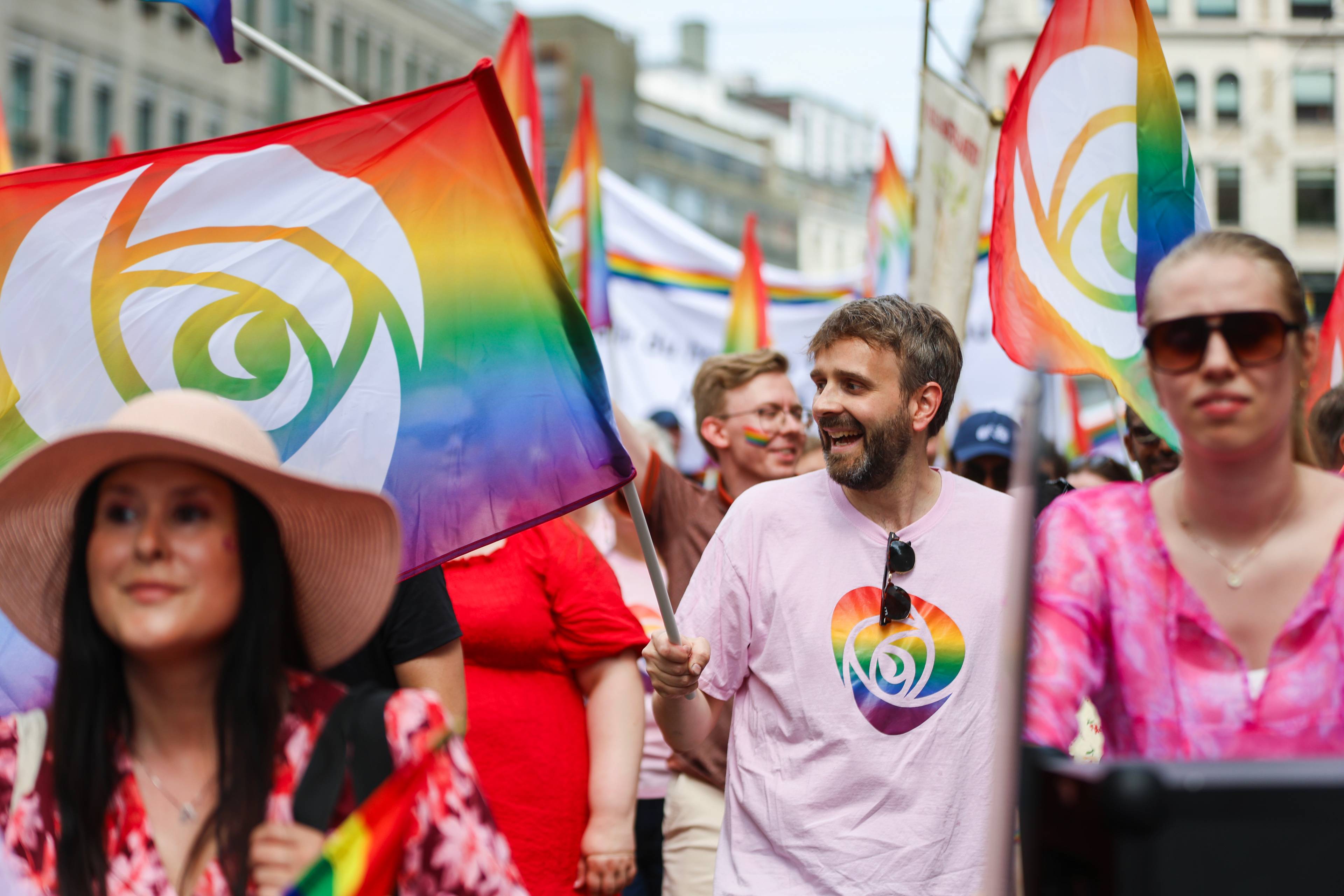 Personer i pride-tog med regnbueflagg med rosemerke. En mann i lyserosa t-skjorte smiler i folkemengden. (Bildebeskrivelsen er laget av en KI-tjeneste)