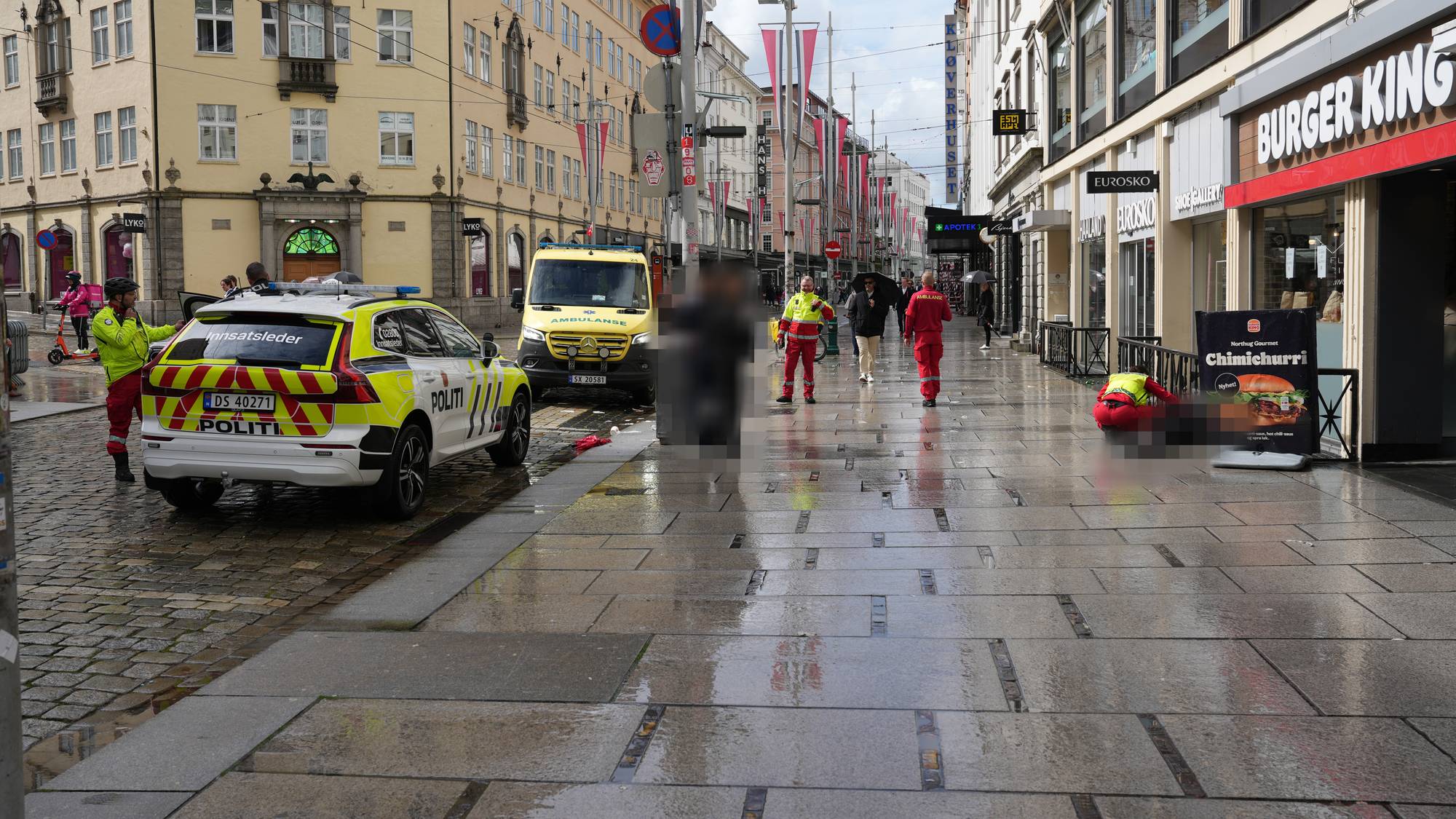 Politi og ambulanse i Strandgaten i Bergen sentrum i forbindelse med vold og rus under 17.mai-feiringen. To ambulansearbeidere sitter på huk ved en dresskledd mann som ligger på fortauet og trenger helsehjelp. 