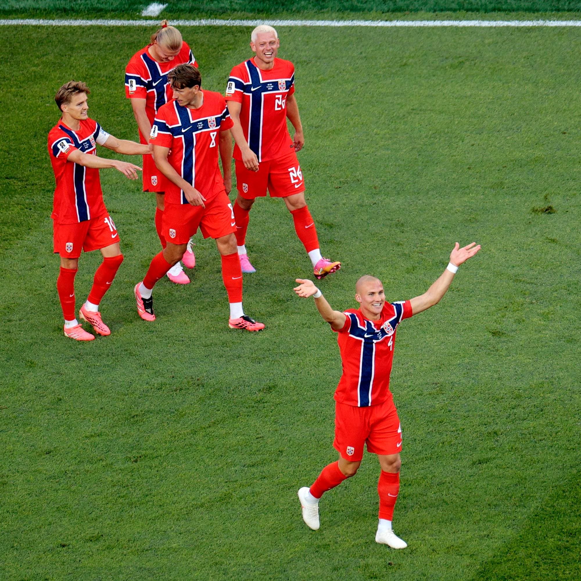 Fotballspillere i røde drakter feirer på en gressmatte. En spiller i forgrunnen med armene ut til siden. Flere andre spillere står bak og ser mot ham. Bildet er tatt fra siden. (Bildebeskrivelsen er laget av en KI-tjeneste)