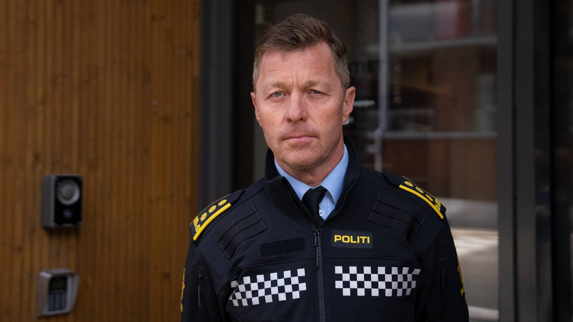 Portrett av en mann i politiuniform. Han har kort, lyst hår og blå øyne. Mannen står foran en bygning med en tredør og et glassparti.