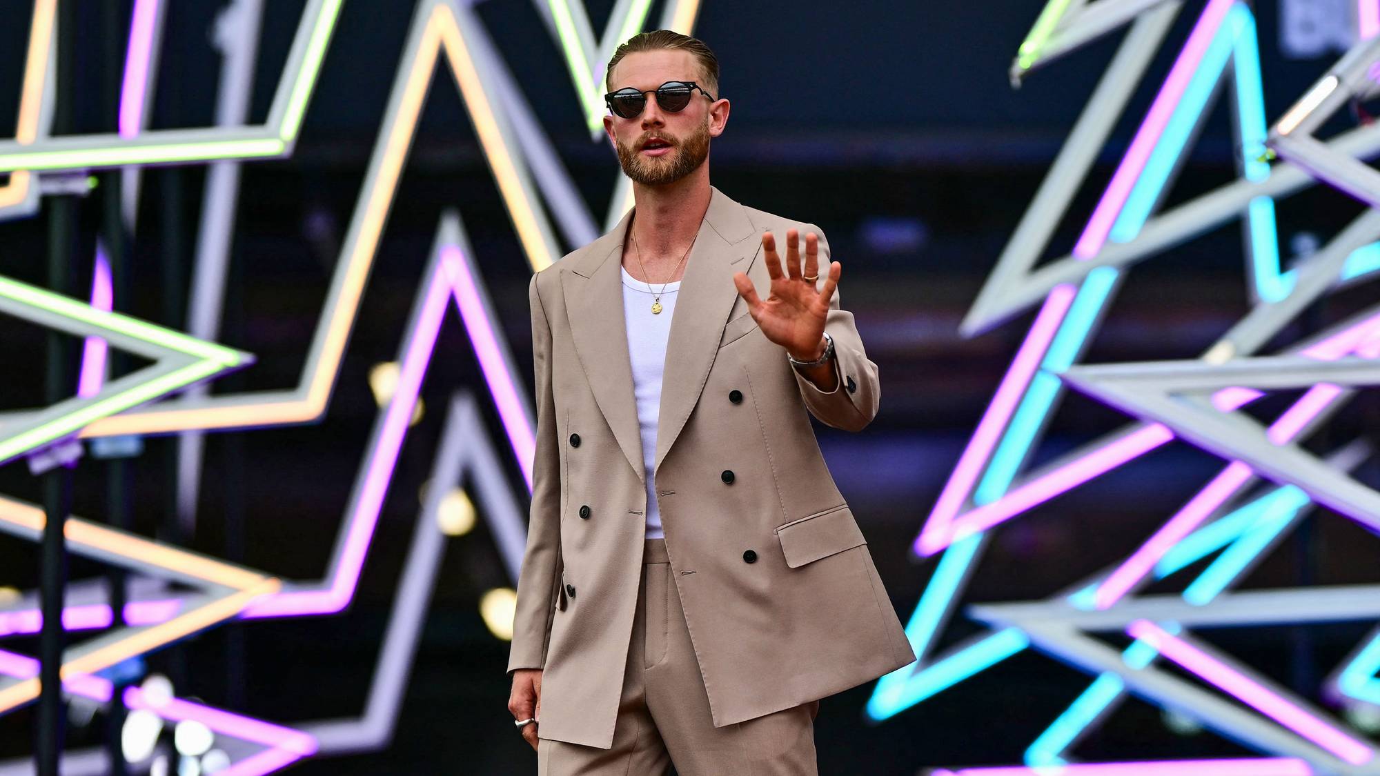 Mann i beige dress hilser med hånden foran en dekorasjon med lysende neonstjerner. (Bildebeskrivelsen er laget av en KI-tjeneste)