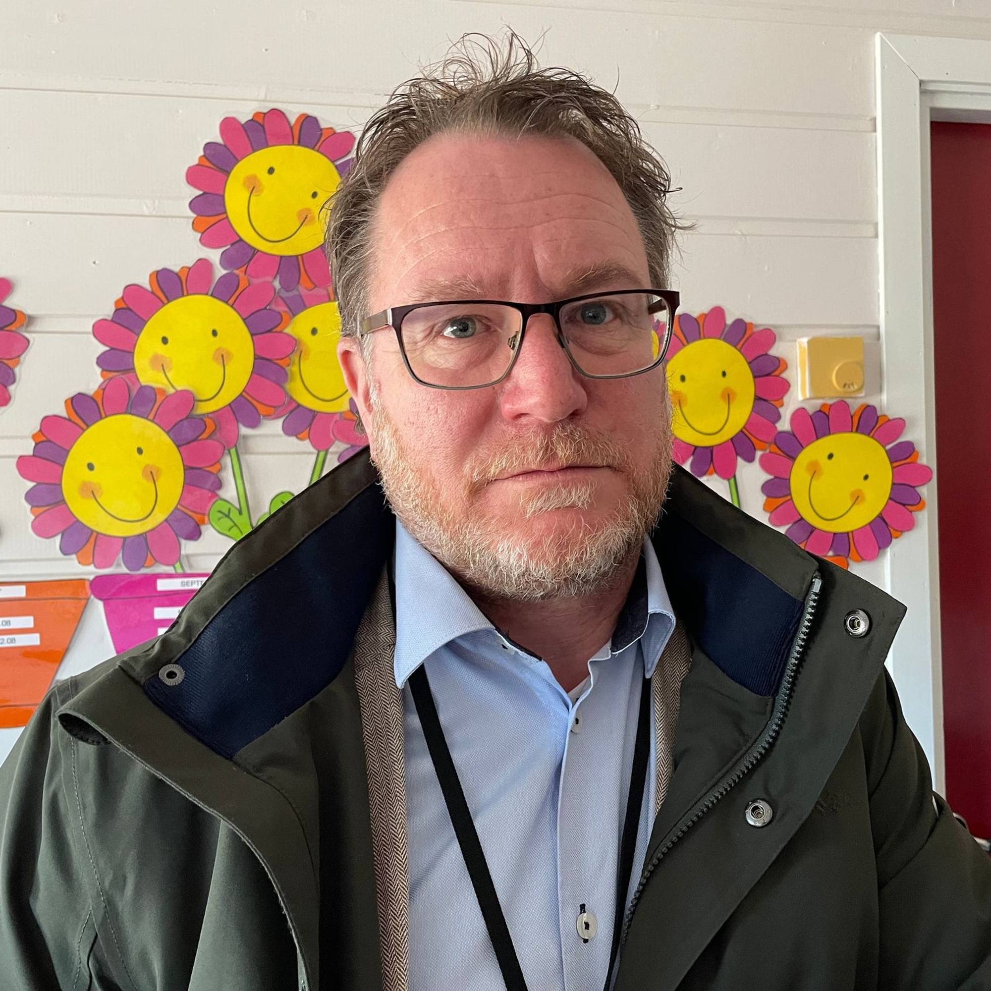 Mann med briller ser inn i kamera. Han er kledd i mørk jakke over lyseblå skjorte. Bak ham er det fargerike bilde av blomster. (Bildebeskrivelsen er laget av en KI-tjeneste)