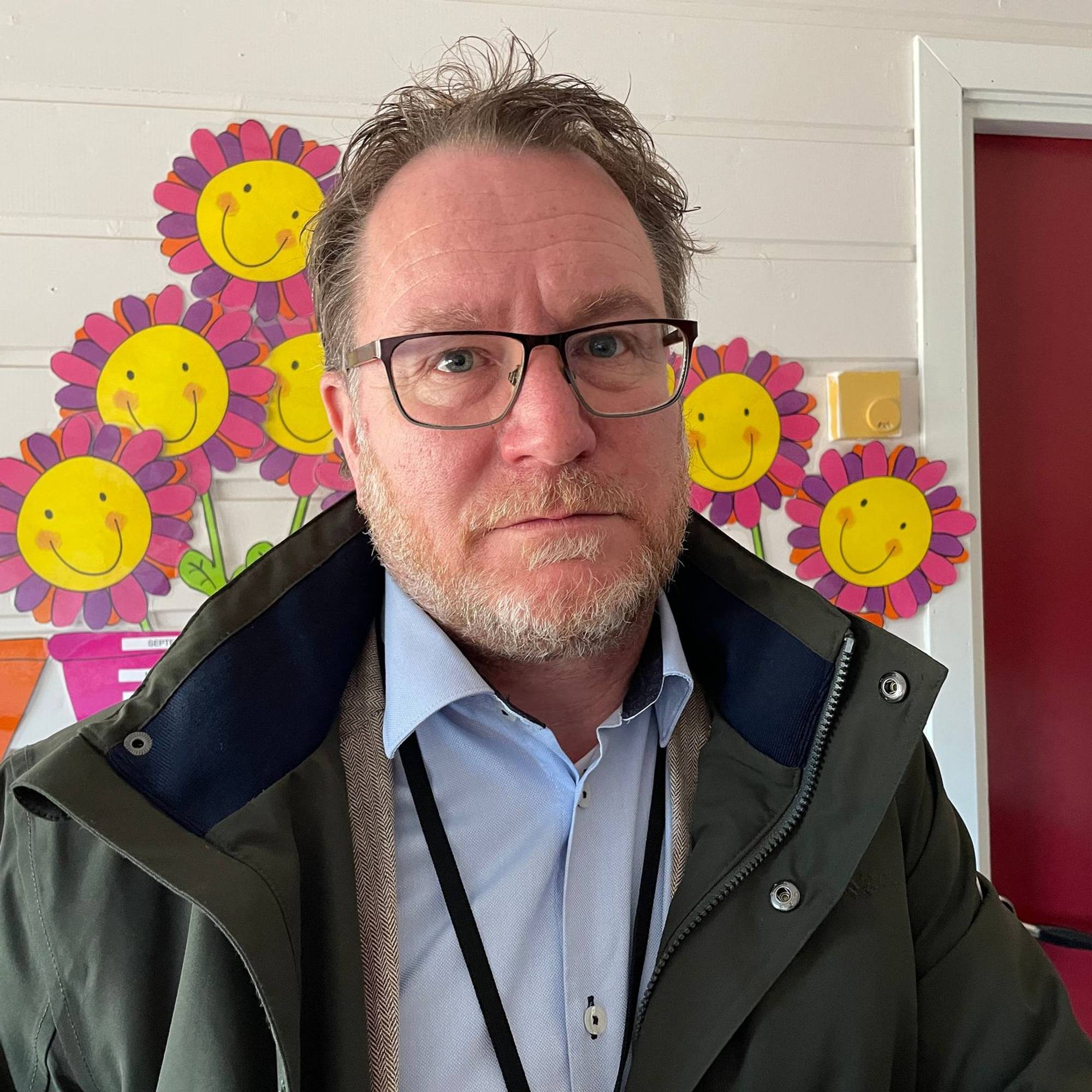 Mann med briller ser inn i kamera. Han er kledd i mørk jakke over lyseblå skjorte. Bak ham er det fargerike bilde av blomster. (Bildebeskrivelsen er laget av en KI-tjeneste)