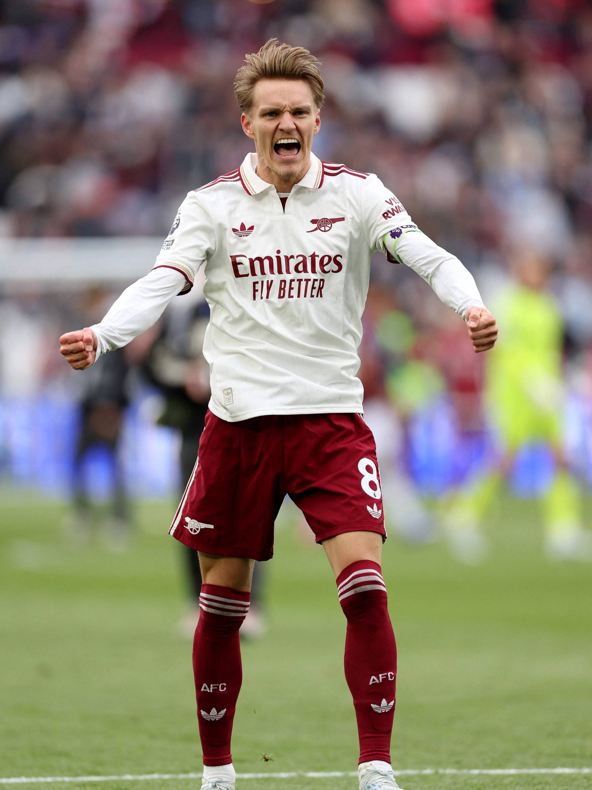 Fotballspilleren Martin Ødegaard jubler under en kamp. Han har på seg Arsenals hvite bortetrøye med maroonfargede detaljer. Spilleren står på en fotballbane, med utydelige tilskuere i bakgrunnen. (Bildebeskrivelsen er laget av en KI-tjeneste)