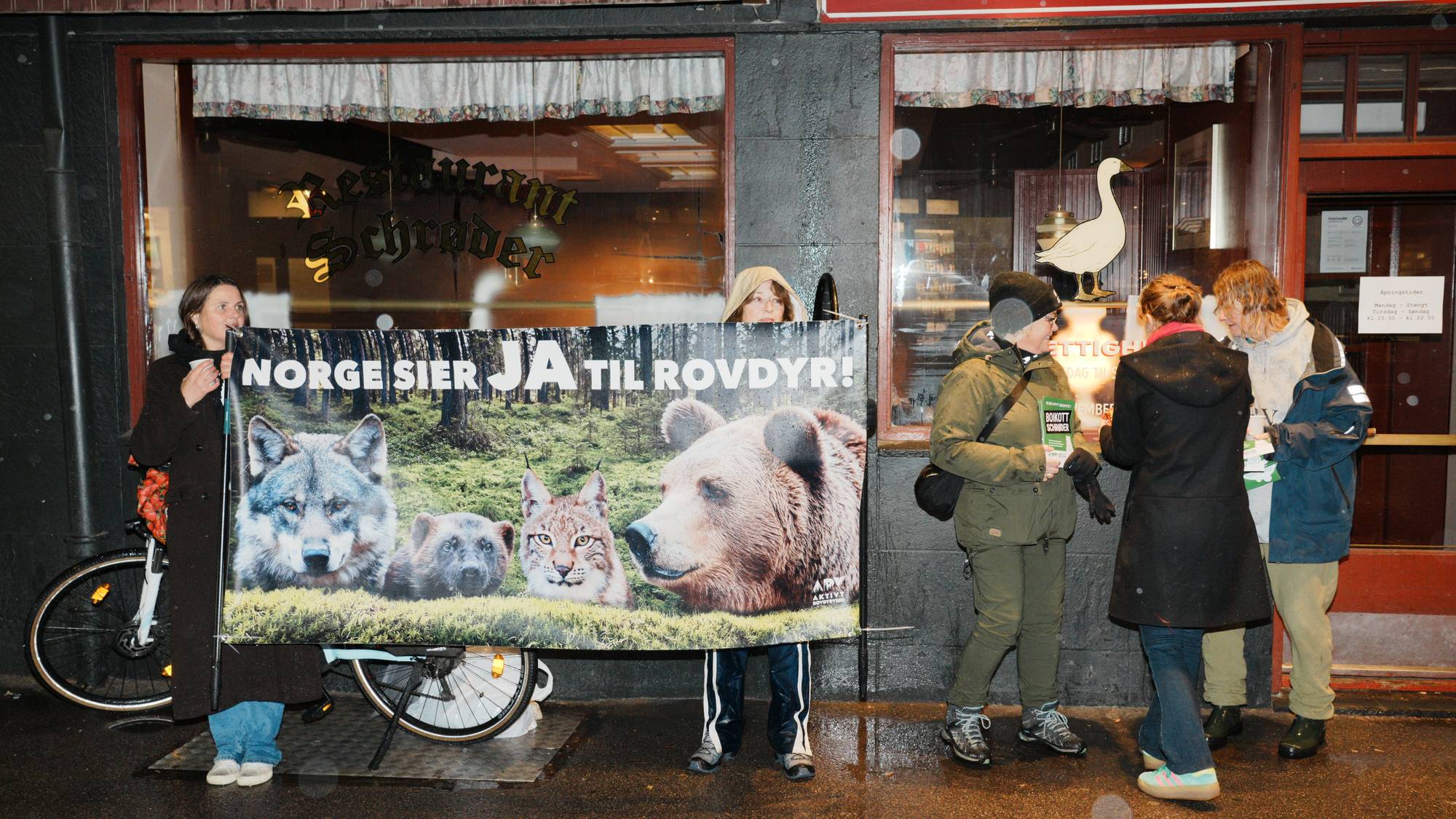 Personer holder en plakat med teksten Norge sier JA til rovdyr utenfor Restaurant Schrøder. (Bildebeskrivelsen er laget av en KI-tjeneste)