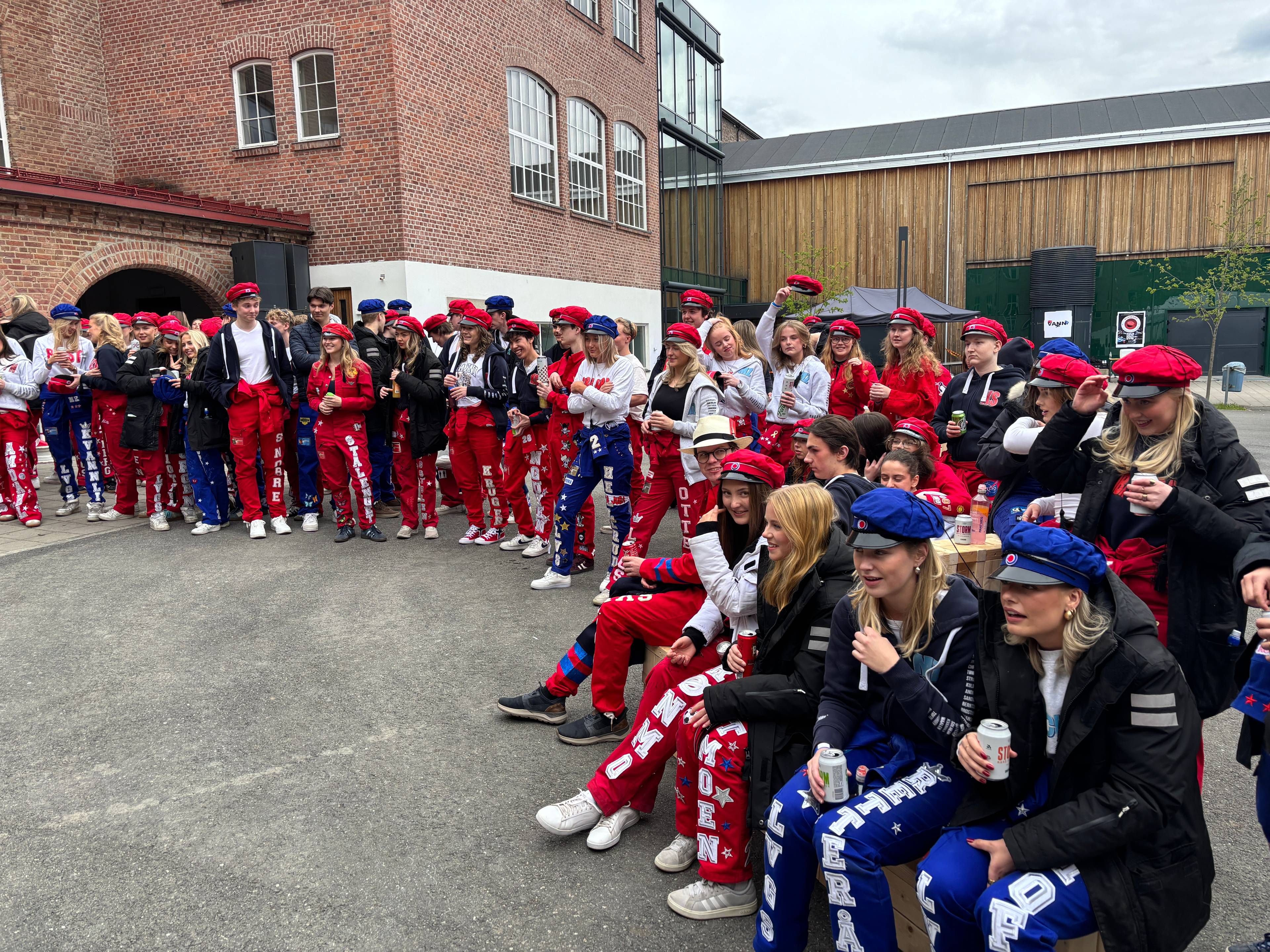 En gruppe ungdommer i røde og blå russeklær sitter og står sammen ute. Mange har på seg russeluer.