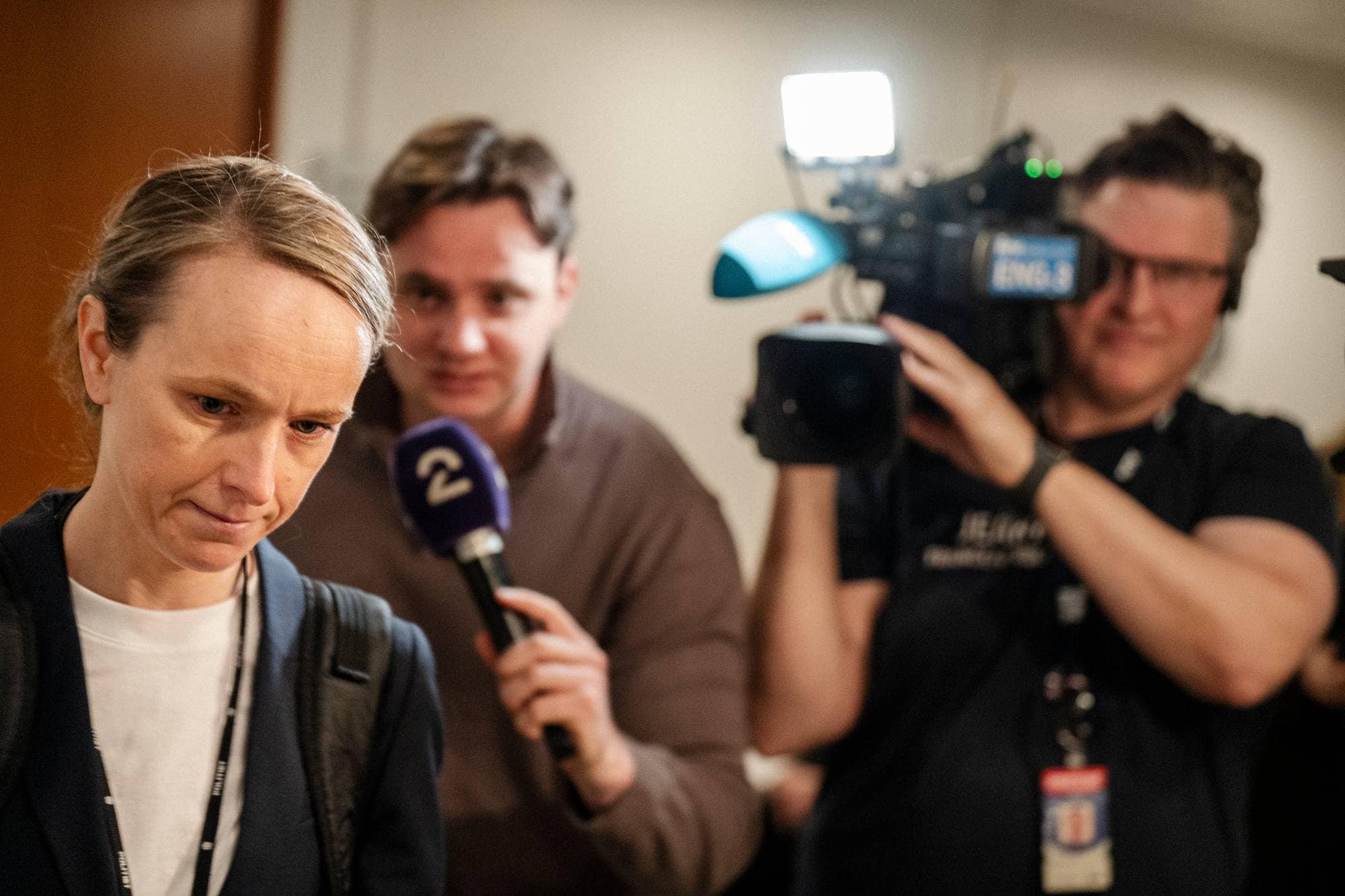 Kvinne med konsentrert ansiktsuttrykk ser ned. I bakgrunnen en fotograf og en journalist med mikrofon