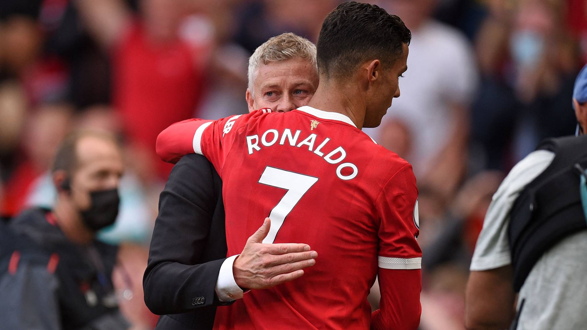 Cristiano Ronaldo omfavnes av Ole Gunnar Solskjær på en fotballbane. Ronaldo bærer en rød drakt med nummer 7. Tilskuere i bakgrunnen har på seg røde klær. (Bildebeskrivelsen er laget av en KI-tjeneste)