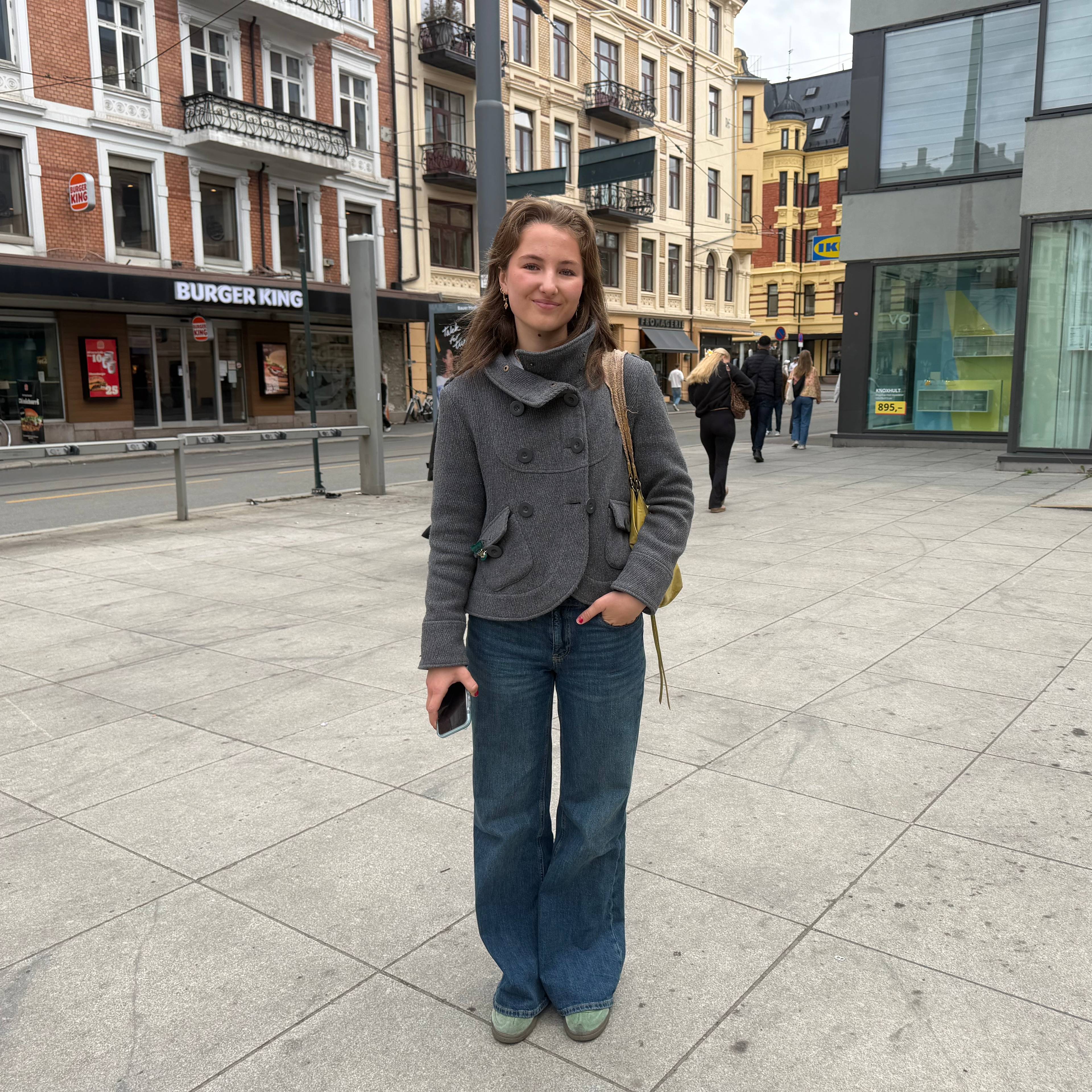 En ung kvinne står på fortau. Hun har en grå jakke, blå jeans og en gul veske. I bakgrunnen sees bygninger med butikker. En av butikkene er IKEA. (Bildebeskrivelsen er laget av en KI-tjeneste)