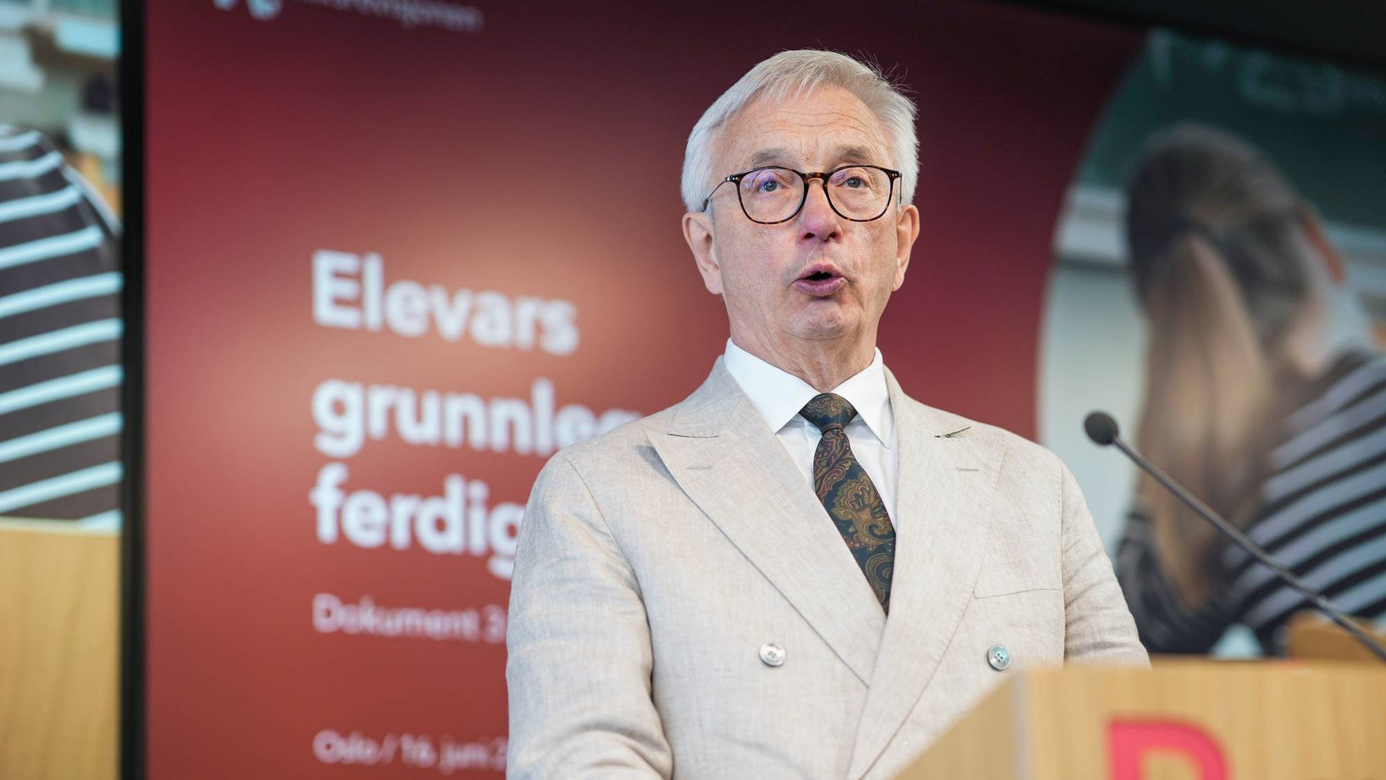 En mann i dress snakker ved et podium. Bak ham vises en skjerm med teksten "Elevars grunnleggjerede ferdigheter". En mikrofon står foran podiumet. Det er også en person i bakgrunnen med en stripete genser. 