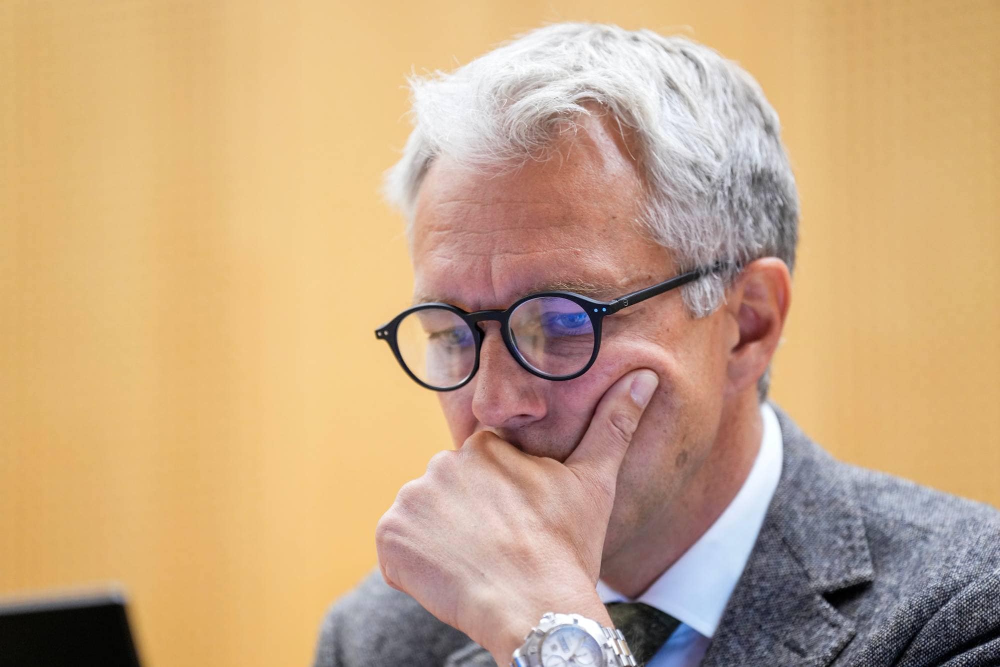 Mann med briller og grått hår holder hånden mot ansiktet foran en lys vegg. (Bildebeskrivelsen er laget av en KI-tjeneste)