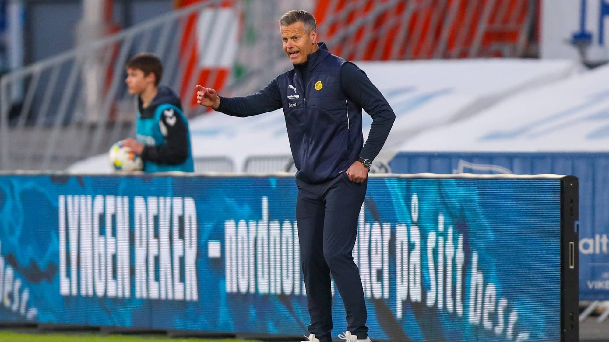 Fotballtrener snakker til sitt lag. Han står på kunstgresset med en reklameskjerm i bakgrunnen. En gutt med fotball står i bakgrunnen. (Bildebeskrivelsen er laget av en KI-tjeneste)