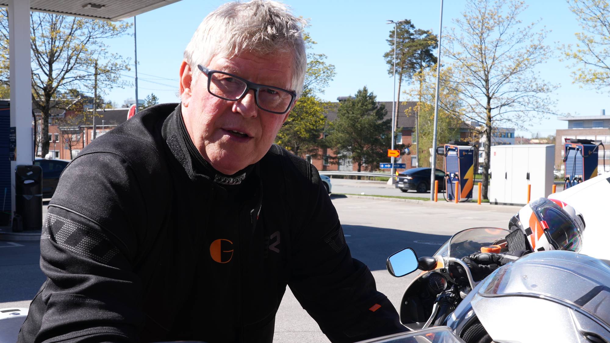 En mann med briller og motorsykkelklær ser mot kamera. I bakgrunnen er en ladestasjon og flere biler. To hjelmer ligger på en motorsykkel. (Bildebeskrivelsen er laget av en KI-tjeneste)