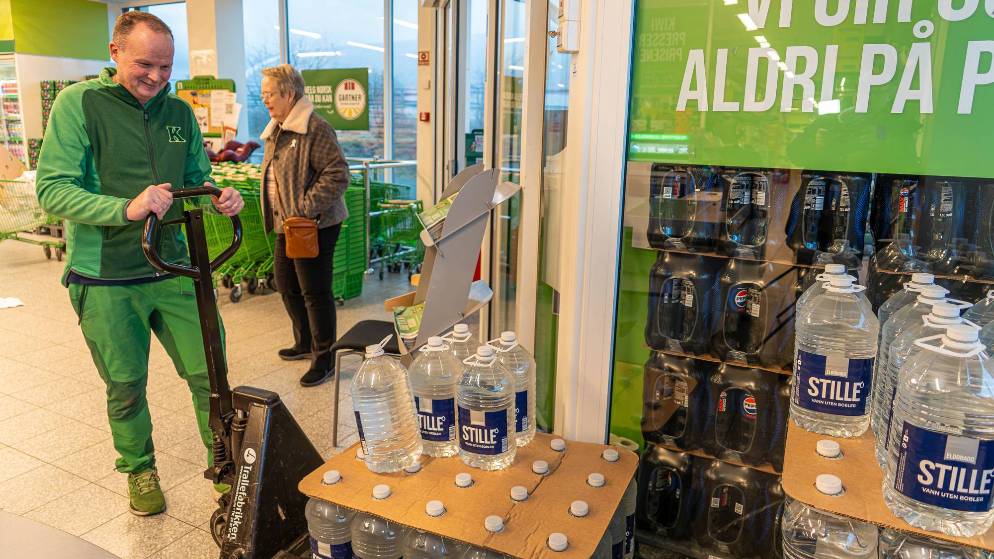 En Kiwi-ansatt bruker en jekketralle i en dagligvarebutikk mens en kunde går forbi. (Bildebeskrivelsen er laget av en KI-tjeneste)
