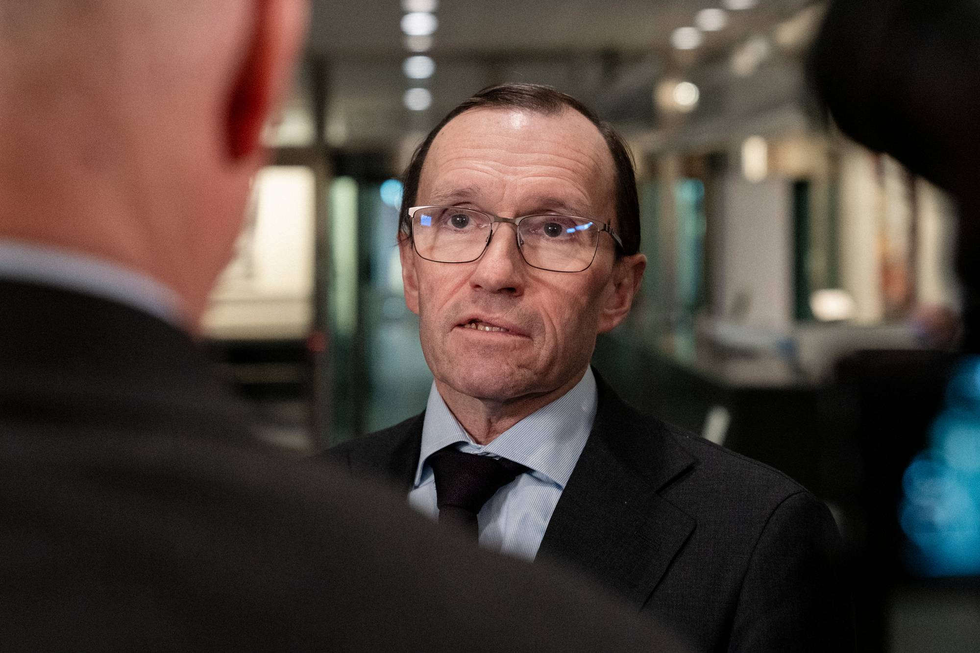 Etter en pressekonferanse står en mann i dress med en nøytral ansiktsuttrykk. Han er omgitt av flere personer, delvis skjult av kameravinkler. (Bildebeskrivelsen er laget av en KI-tjeneste)