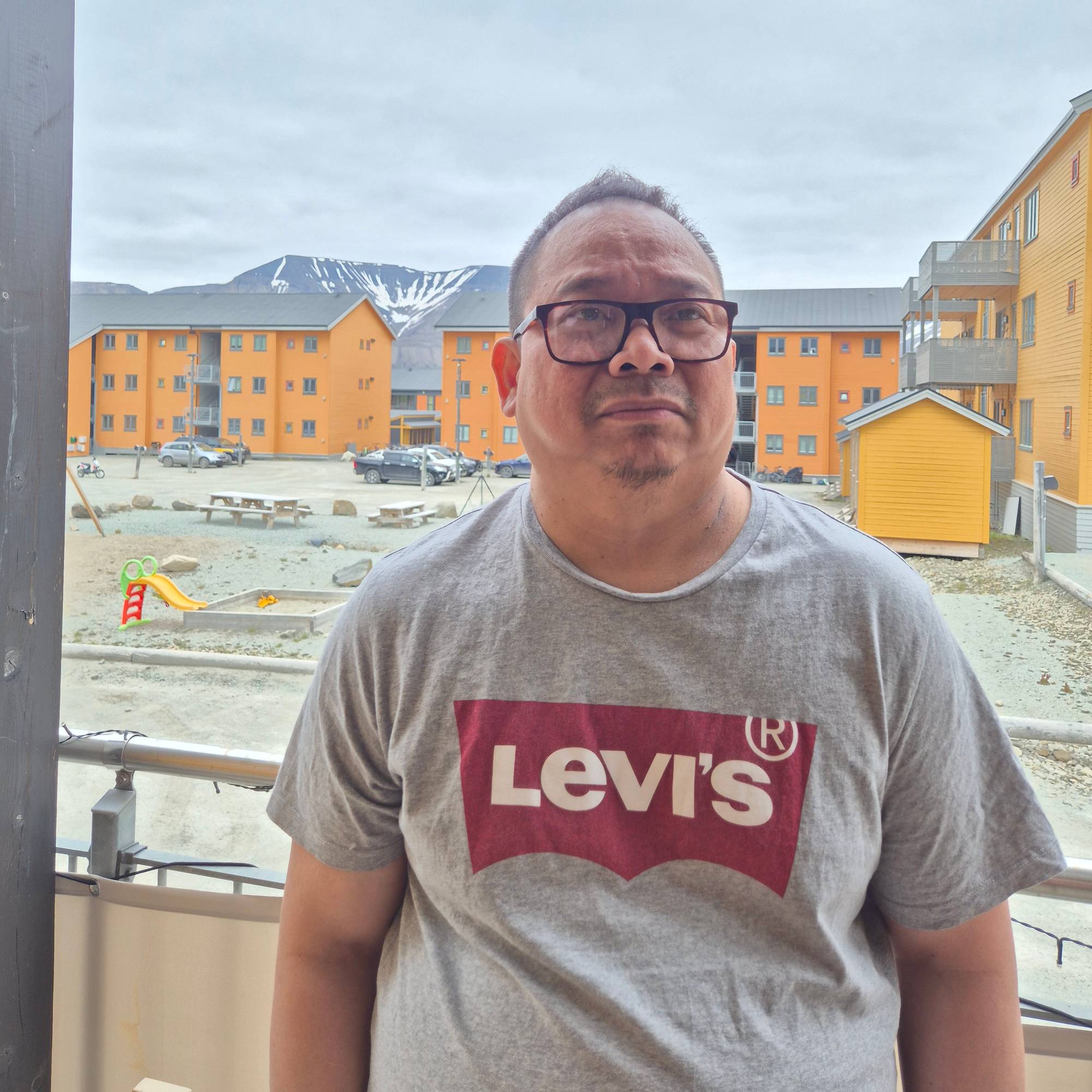 En mann fra Filippinene poserer til et bilde på Svalbard. Han har på seg grå t-skjorte med rød Levis-logo. Mannen har briller og kort mørkt hår. I bakgrunnen er det gule leilighetsbygg, og fjell bak der igjen. 