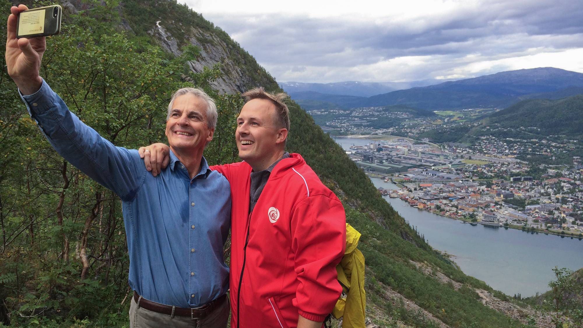 To menn tar en selfie på en fjelltopp. Den ene mannen har en blå skjorte og den andre en rød jakke. I bakgrunnen ser man et fjellandskap med en by og en elv. (Bildebeskrivelsen er laget av en KI-tjeneste)