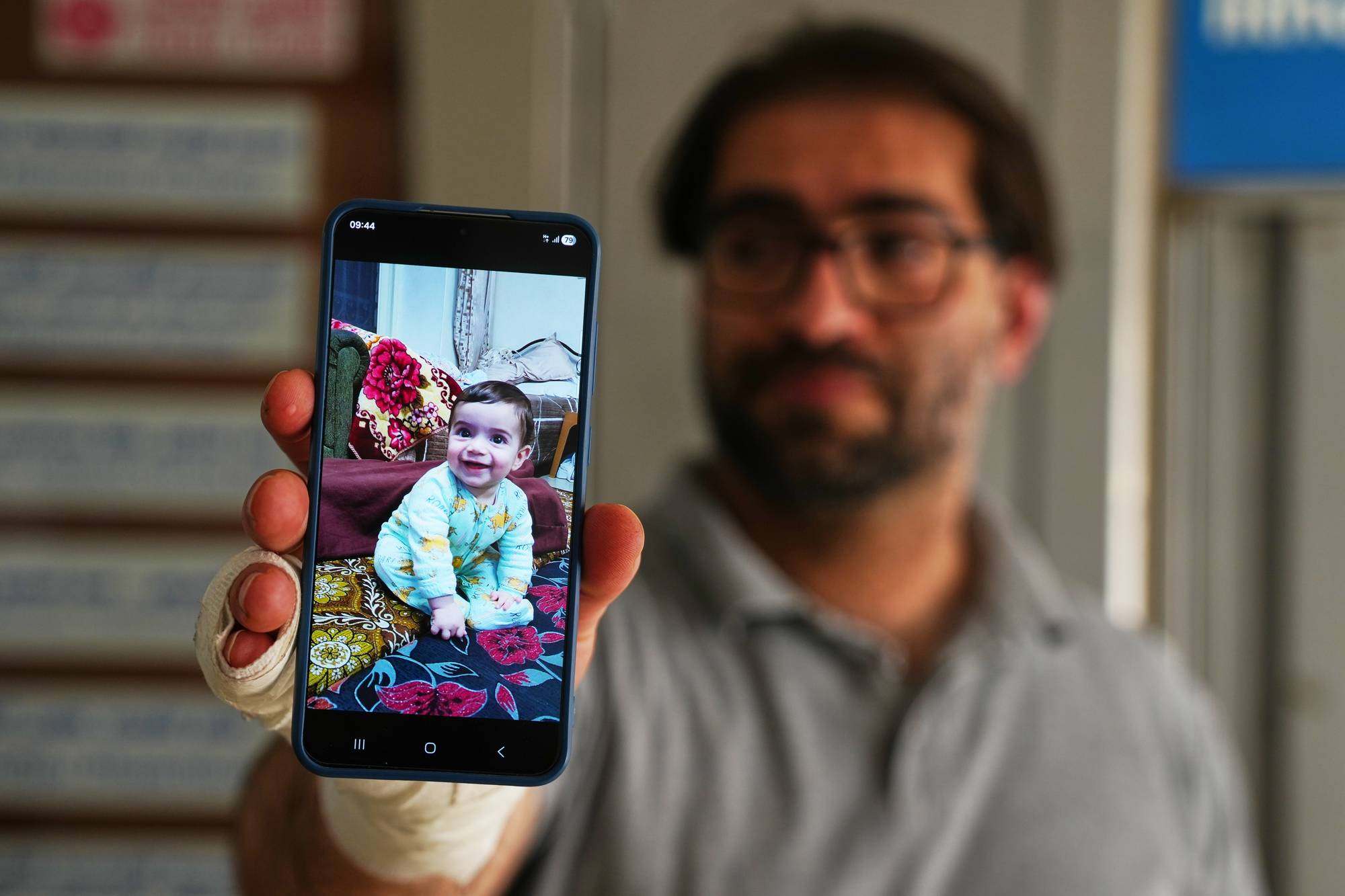 En mann med bandasjert hånd holder opp en mobiltelefon. På mobilskjermen vises et bilde av en baby. Mannen har briller.