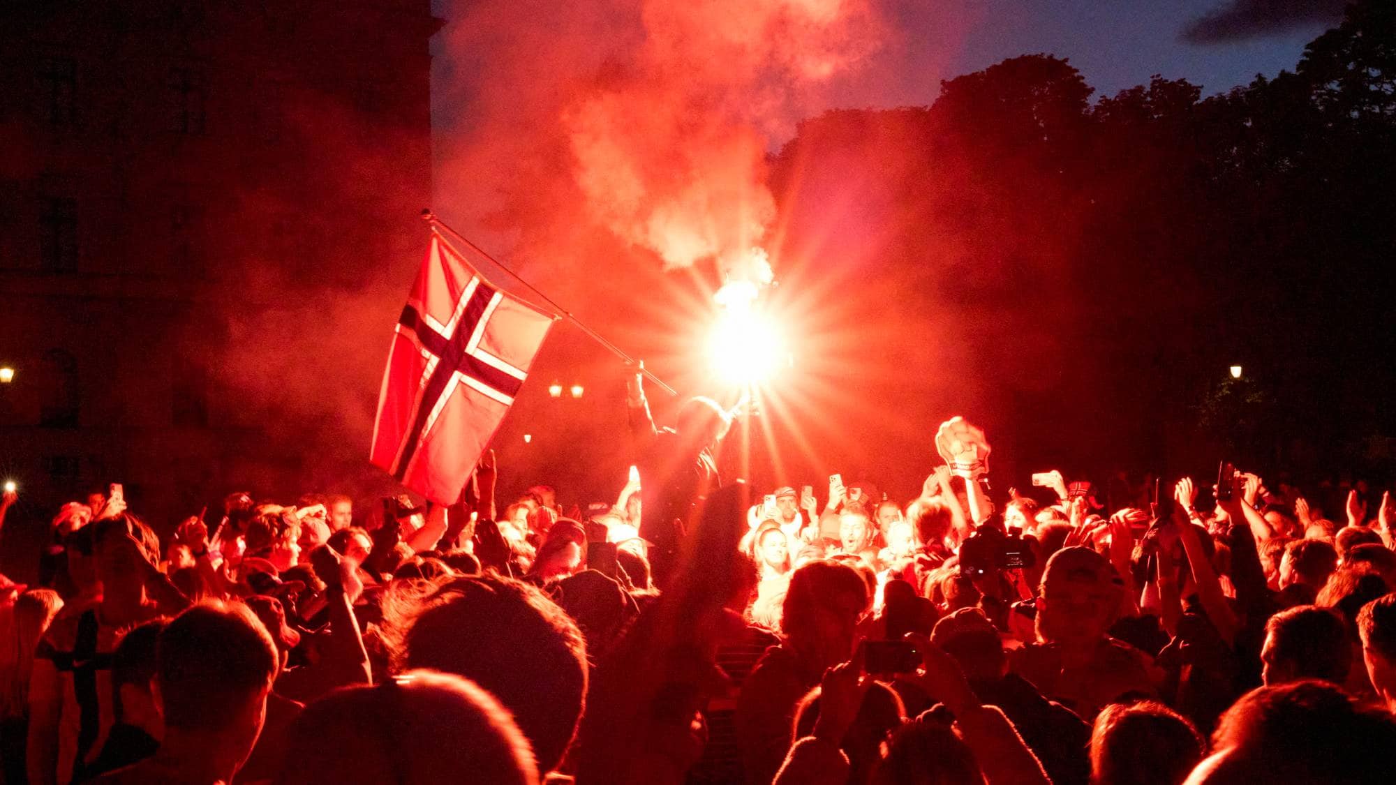 Person holder norsk flagg og lysbluss foran en folkemengde i skumringen. (Bildebeskrivelsen er laget av en KI-tjeneste)