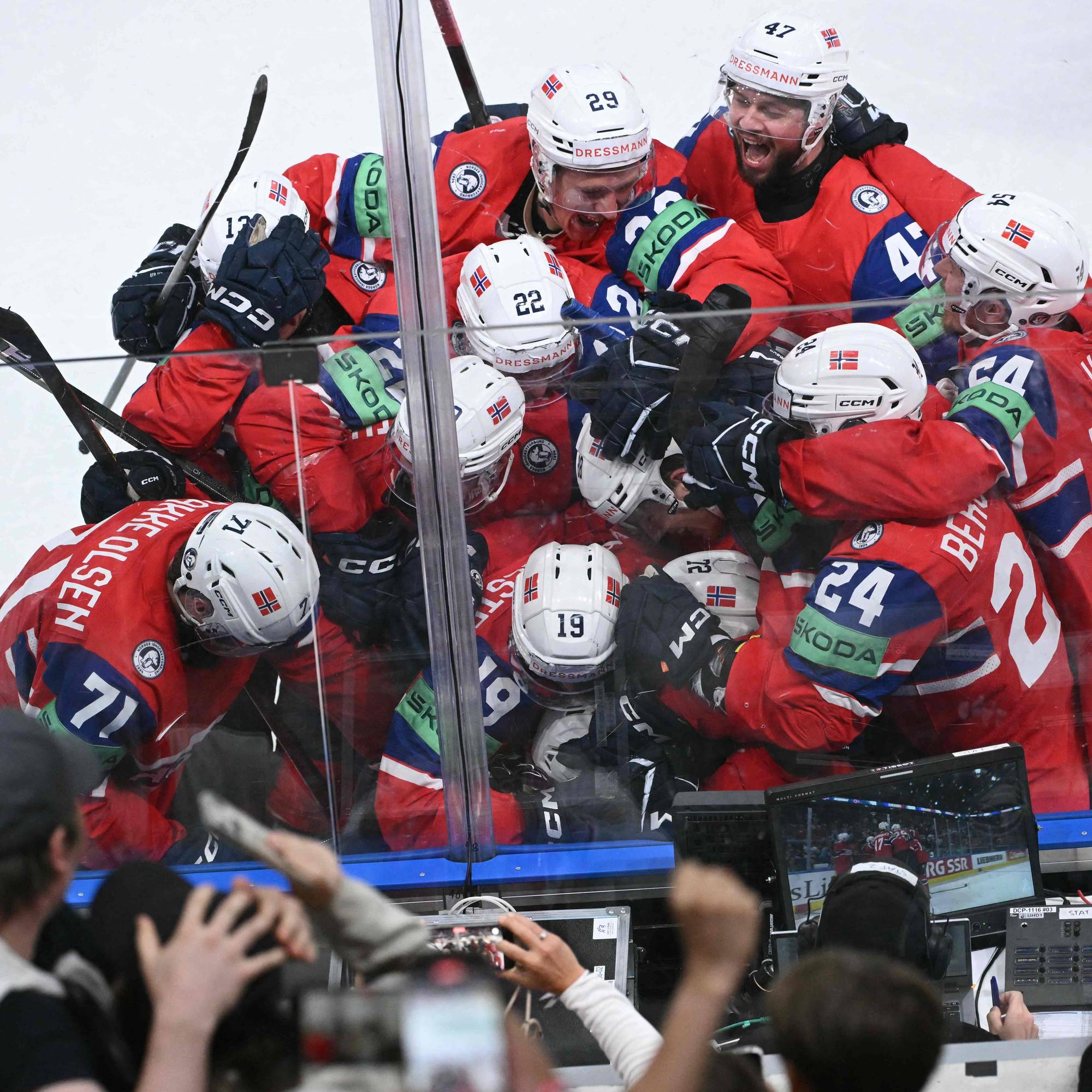 Ishockeylandslaget jubler sammen på isen. Spillerne har på seg røde landslagsdrakter med nummer og navn. En skjerm viser et klipp fra kampen. I forgrunnen ser man publikum som holder opp mobiler for å ta bilde. (Bildebeskrivelsen er laget av en KI-tjeneste)