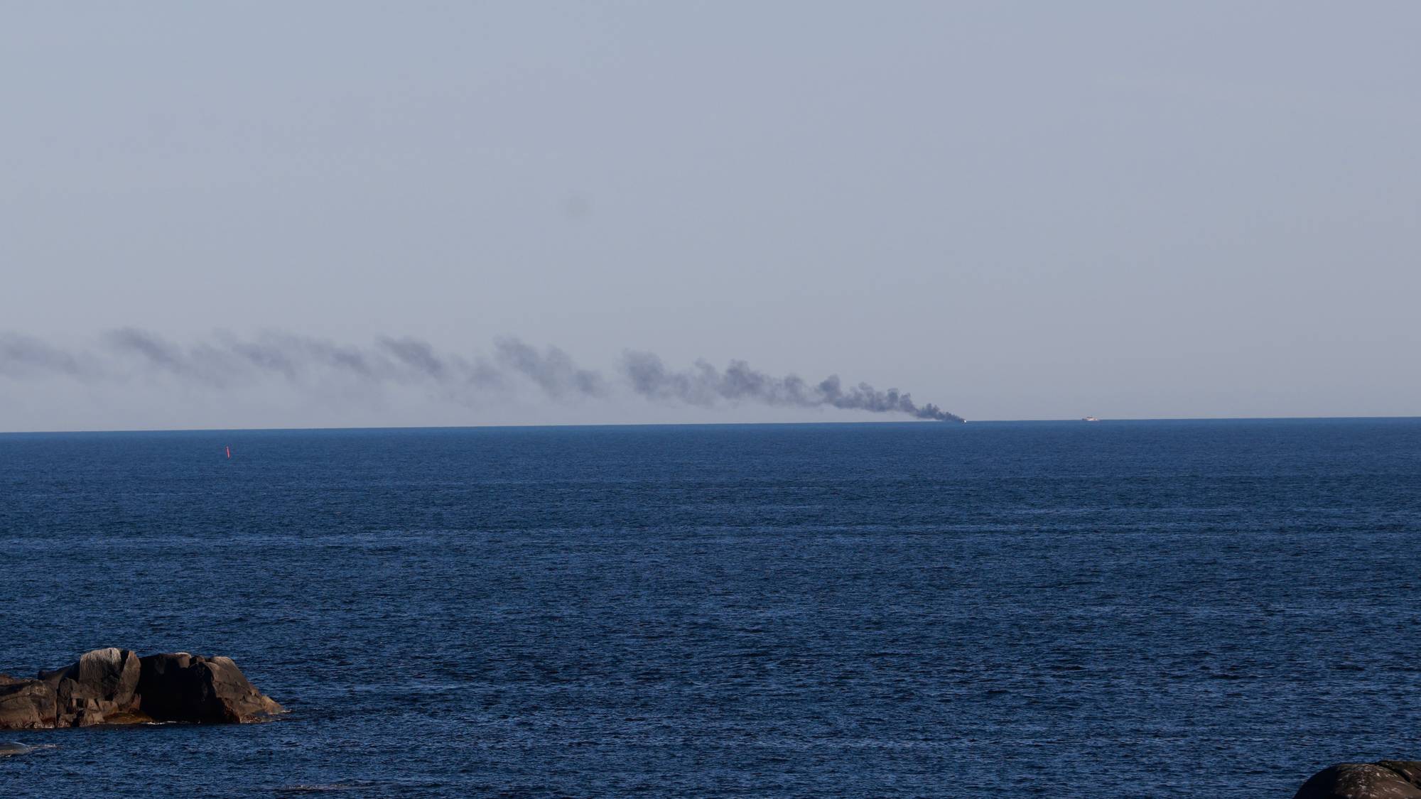En røyksøyle stiger opp fra havet langt ute. Det er flere båter på det dype blå vannet. Det er noen steiner i forgrunnen. (Bildebeskrivelsen er laget av en KI-tjeneste)