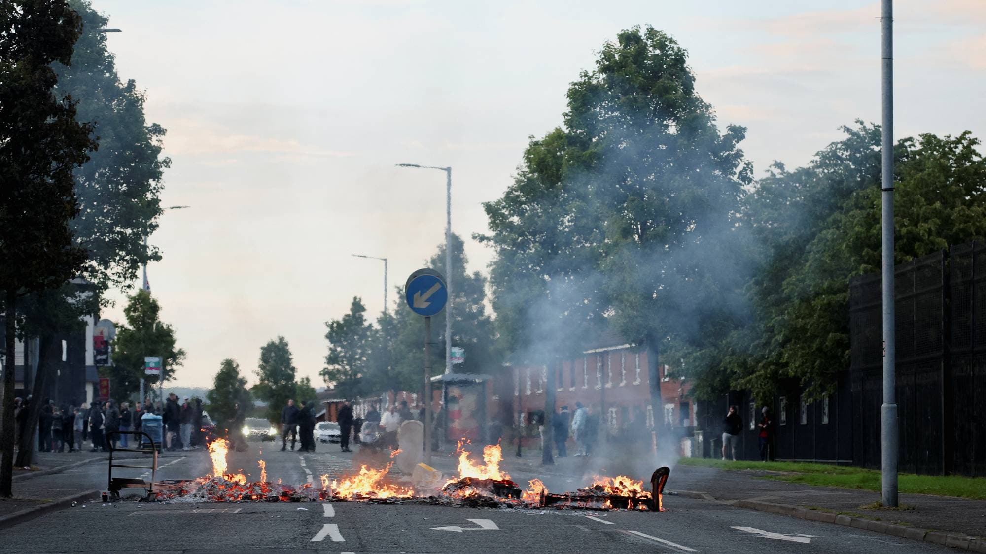 En gate er blokkert av brennende gjenstander og røyk. Flere personer står på fortauet og ser på. Bildet er tatt i et boligområde med mursteinshus og trær. (Bildebeskrivelsen er laget av en KI-tjeneste)