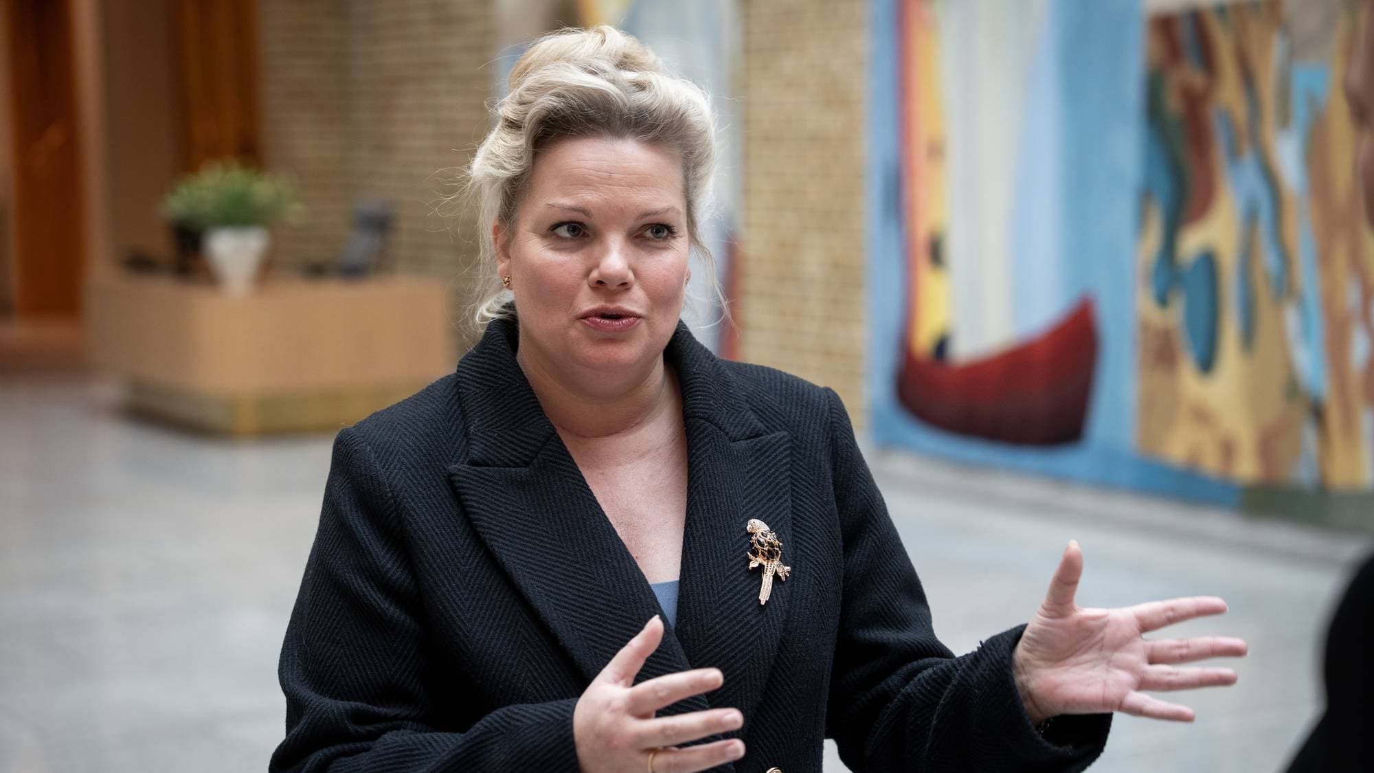 En kvinne med blondt hår i oppsetning snakker og gestikulerer foran en abstrakt maleri. Hun har på seg en mørk dressjakke med en brosje formet som en fugl. Bak henne ses et interiør med planter og vegger. (Bildebeskrivelsen er laget av en KI-tjeneste)