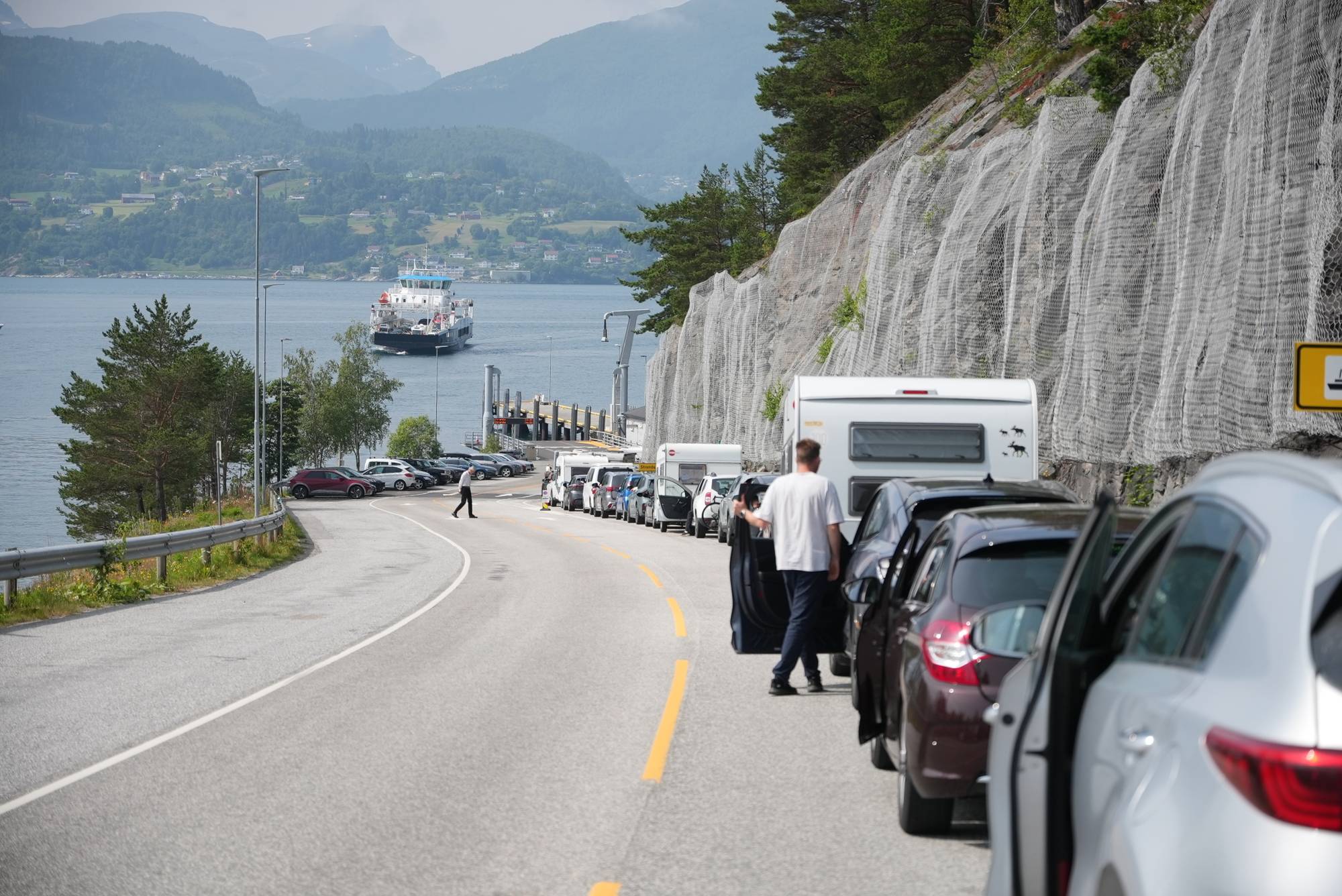 Bilister venter i kø foran en fergekai ved en vei langs en fjellvegg. (Bildebeskrivelsen er laget av en KI-tjeneste)