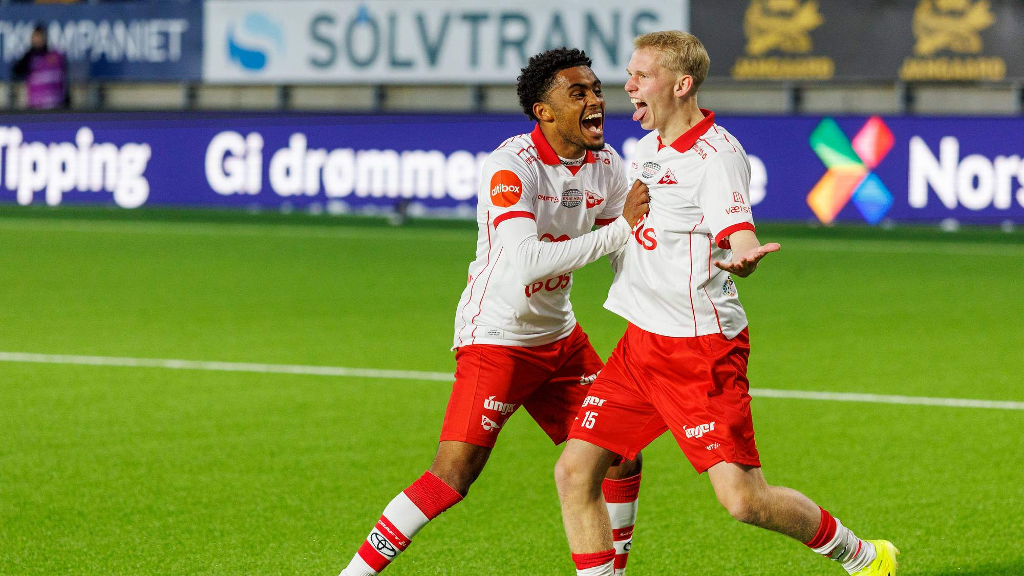 To fotballspillere i hvite og røde drakter jubler på en grønn gressmatte. De er omringet av reklameskilt på en idrettsarena. Spilleren til venstre har tungen ute og holder den andre spilleren i armen. Spilleren til høyre ser opp og har hendene ut til siden. (Bildebeskrivelsen er laget av en KI-tjeneste)