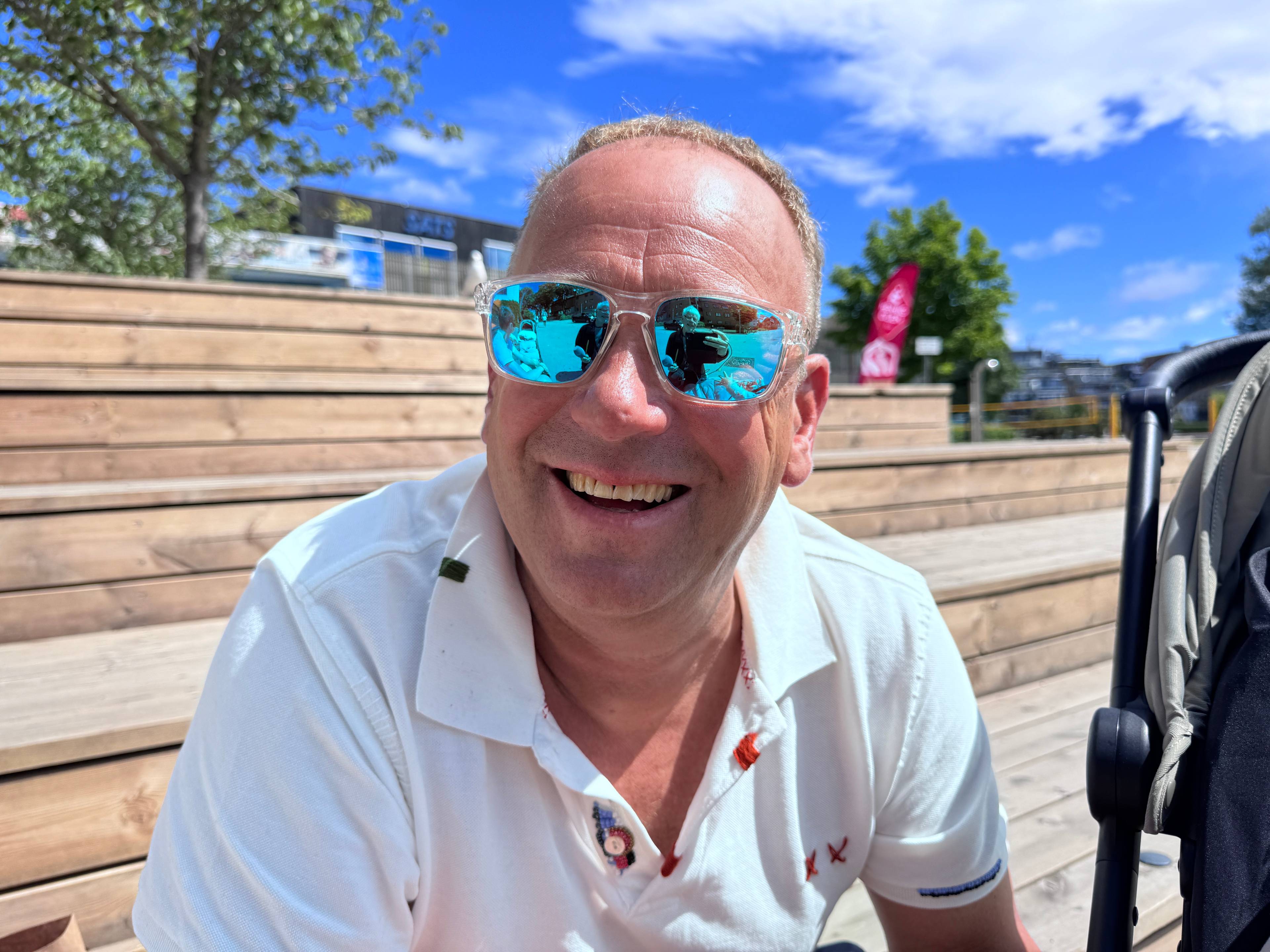 En mann med hvit skjorte og solbriller smiler til kamera. Han sitter på en trapp.